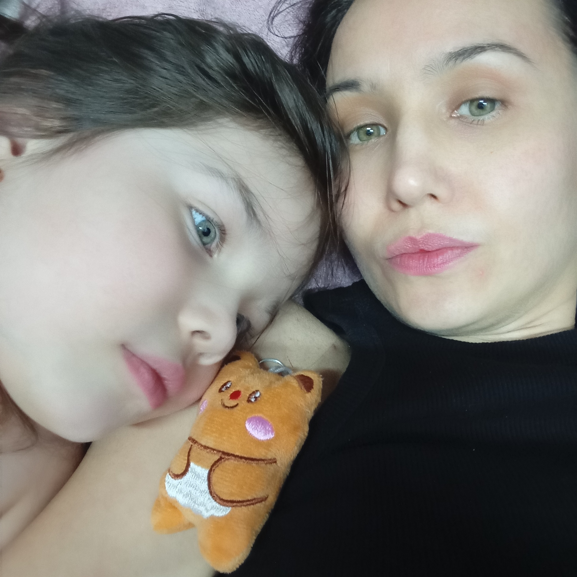 Two people, a child and an adult, are close together. The child has fair skin, blue eyes, and dark hair. The adult, with fair skin and green eyes, wears a black top. A plush toy depicting an animal, brown with white details, is visible near the child. The setting appears to be an indoor environment. There is no information that connects the image to the city of Pervomaysky, Russia.