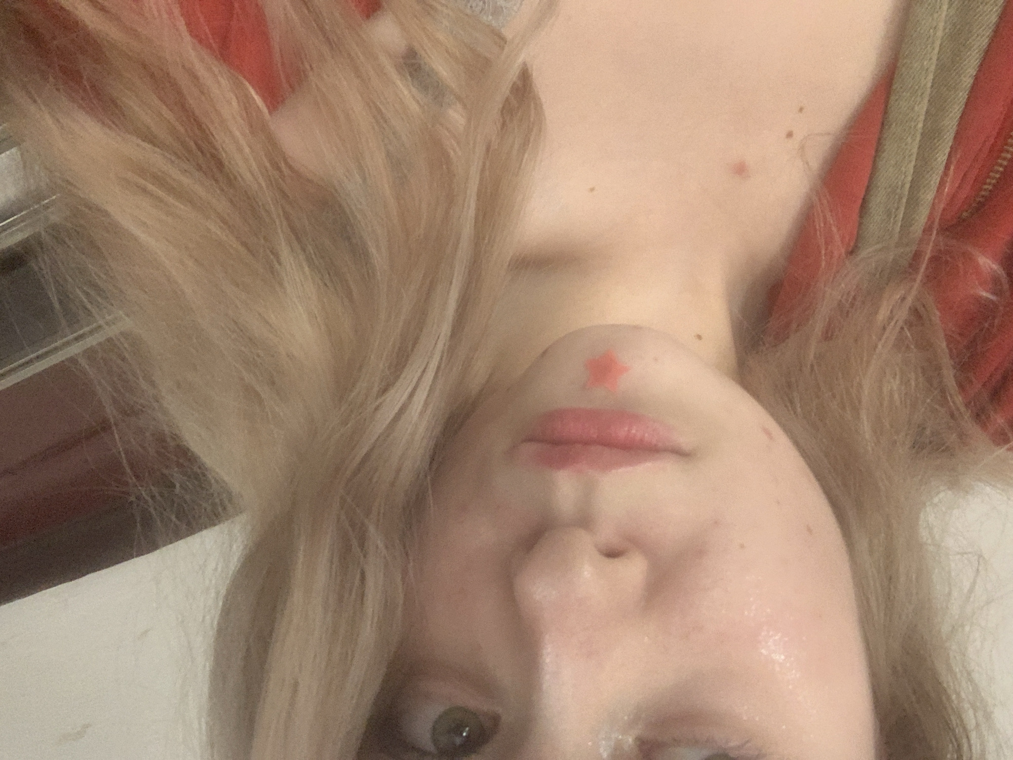 The image displays a close-up, inverted portrait of a person, likely a young woman, with light-colored (blonde or light brown) hair. Her face is upside down, with her chin positioned towards the top center of the frame and her eye towards the bottom.
**Subjects:** The primary subject is a person, whose face is partially visible, focusing on the chin, mouth, nose, and one eye. Long, wavy, blonde hair frames the face and cascades towards the top and left of the image. On the right side, a red garment with a visible zipper and a khaki or olive-green strap is seen, possibly a jacket or top.
**Activity or event taking place:** The person is looking upwards towards the camera with a direct gaze from her visible eye. It appears to be a selfie or a close-up portrait.
**Notable details:**
- **Orientation:** The entire image is inverted (upside down).
- **Facial Features:** The skin is fair with visible freckles and appears somewhat dewy or oily. Her lips are slightly parted. A distinctive small, red star-shaped sticker or drawing is placed on her chin. Her visible eye is light-colored, possibly green or hazel.
- **Hair:** The hair is voluminous and has a soft, flowing texture.
- **Clothing:** The red garment provides a contrasting color element.
- **Emotion:** The expression is generally neutral or contemplative, with a direct look at the viewer.
- **Scene/Setting:** The background is mostly blurred and indistinct, suggesting an indoor setting without specific location cues.
- **Time of day/Weather:** Not discernible from the indoor lighting.
- **Visible text:** There is no visible text in the image.