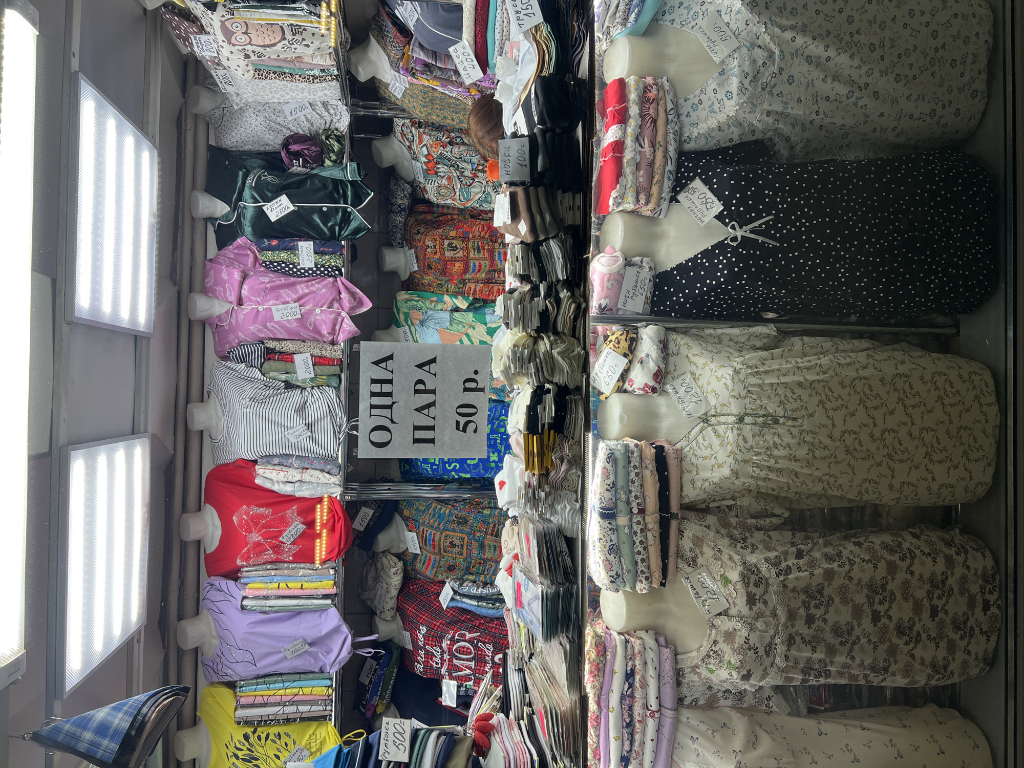 The image shows a retail display, likely in Khimki, Russia, given the Cyrillic script on the signs. Mannequins, primarily headless, display a variety of clothing items, including pajamas, t-shirts, dresses, and socks. Price tags are visible on many items. A sign indicates a promotion: "ODNA PARA 50 p." ("One pair 50 rubles"). The lighting is provided by LED panels. The clothing includes various patterns and colors, from floral dresses to striped shirts and polka-dot tops. The store appears to be well-stocked, and the merchandise is arranged in a dense, vertical manner.