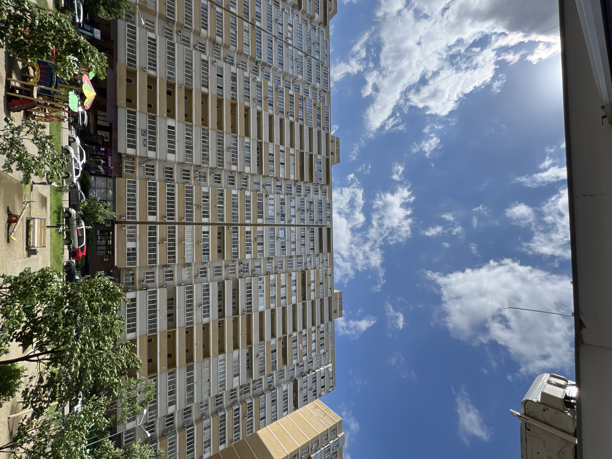 The image shows a tall apartment building in Krasnodar, Russia, viewed from below at an angle. The building has a repetitive pattern of windows and beige-colored balconies. To the left of the building, there are trees, a playground, and parked cars along a street. The sky is blue with scattered white clouds. An air conditioning unit is visible in the lower right corner of the image, attached to a wall.