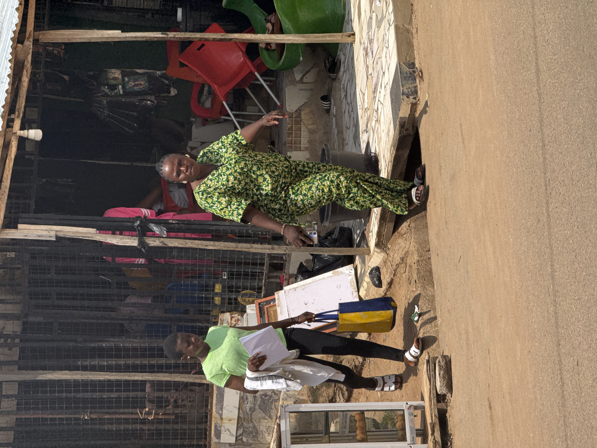 Two women are walking on a dirt path next to a street in Abuja, Nigeria. The woman on the left is wearing a lime green t-shirt and black pants, and carrying a white shirt and a blue and yellow striped bag. The woman on the right is wearing a green and yellow patterned outfit and appears to be walking from a building. The building behind them has a corrugated metal roof and a dark interior with various items displayed. Red chairs are visible inside and outside the building. The ground is sandy and dusty, with scattered debris and a black trash bag. There are also some plants and a glass display case with baked goods on the left side of the image.