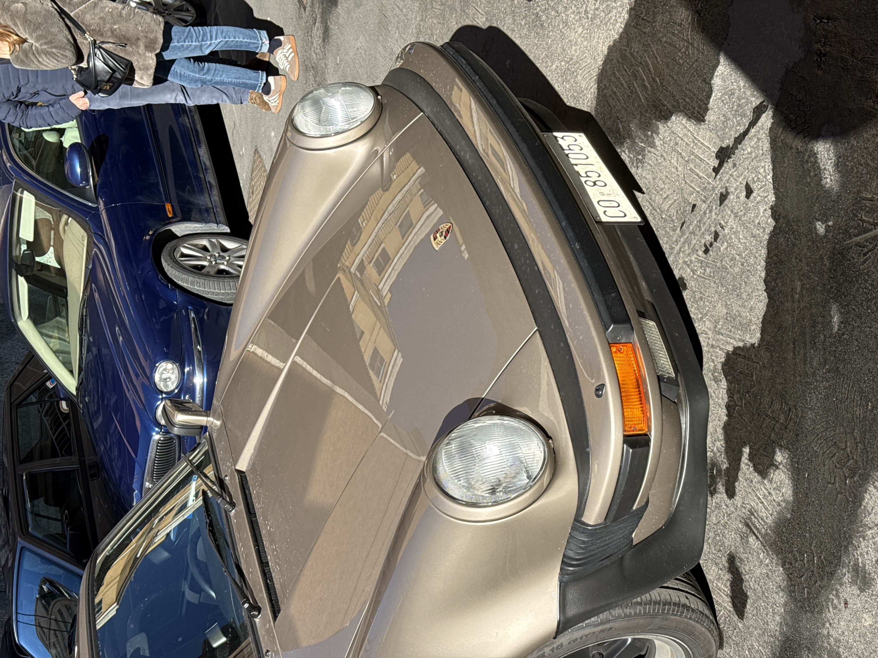 The scene depicts a street in Milan, Italy, featuring three vehicles and two partially visible individuals under direct sunlight, casting distinct shadows on the uneven concrete ground.
In the foreground, a beige-gold Porsche 911, likely a G-series model, is parked facing forward, occupying the majority of the frame. Its front features prominent round headlights, a black rubber bumper with accordion-style bumperettes on the left side, and an integrated rectangular orange turn signal on the right. A Porsche crest is visible on the center of the hood, which shows reflections of a yellow-toned building facade and light dusting. The Italian license plate, "CO 851053," is affixed to the front bumper. The car's front left tire is partially visible.
Behind and to the right of the Porsche, a dark blue Jaguar X-Type sedan is parked. Its chrome grille, headlights, and front passenger side are visible. Reflections of the surrounding architecture are present on its glossy surface.
Two individuals stand adjacent to the front passenger side of the blue Jaguar. The person on the left is a male, wearing a dark blue puffer jacket, light blue jeans, and brown lace-up shoes with white soles. His right hand is in his pocket. The person on the right is a female, wearing a brown faux fur coat, light blue jeans, and brown lace-up shoes similar in style to the male's. She carries a black shoulder bag with a gold-colored clasp across her body. Both individuals' lower legs and torsos are visible, and they appear to be standing still. No direct interaction between them is discernible.