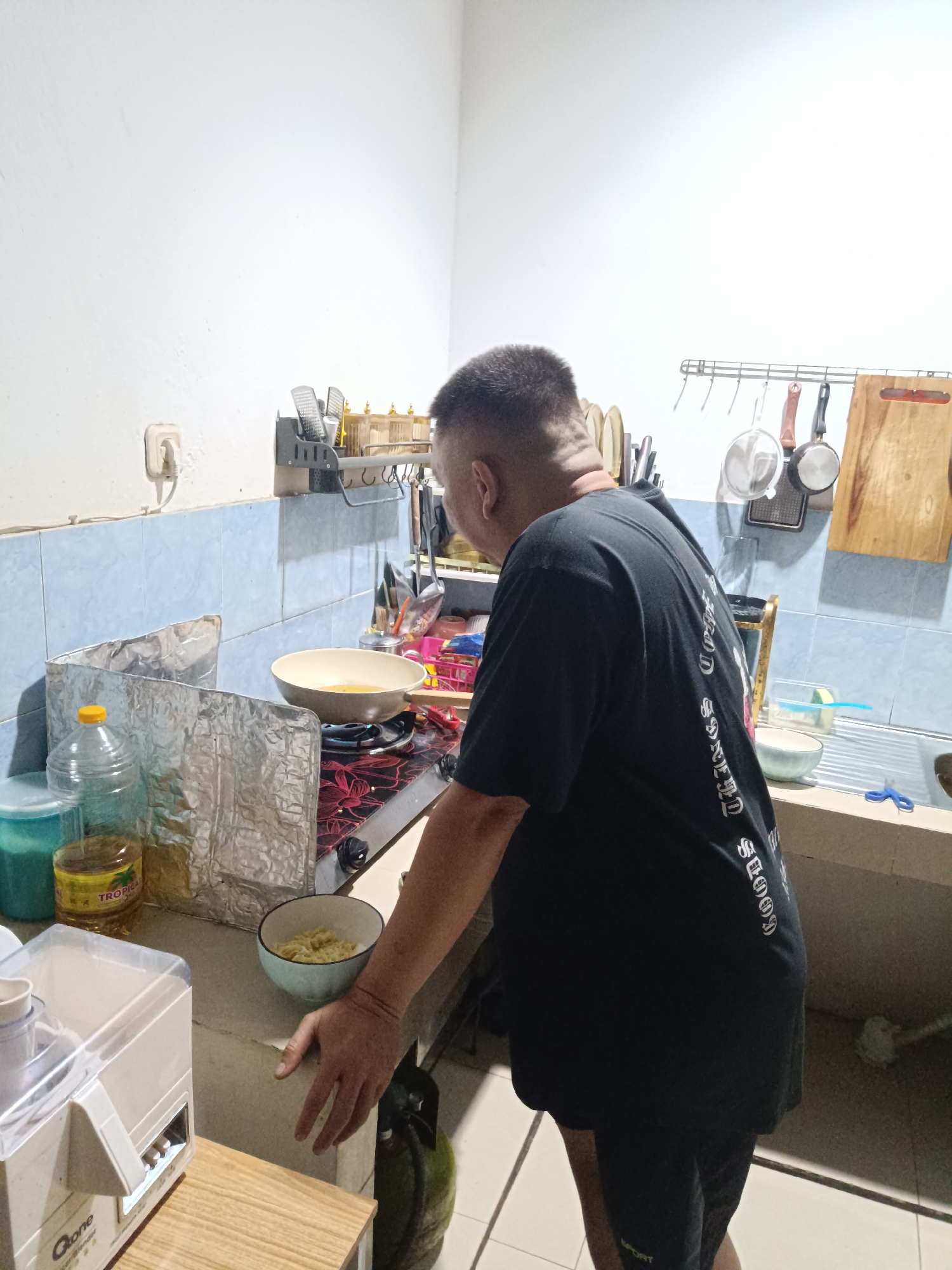 The media file shows a man standing in a kitchen, seemingly cooking. He is wearing a black t-shirt with white text on the back. The kitchen appears to be in a home, with a gas stove, a sink, and various cooking utensils and ingredients visible. The man is facing away from the camera, towards the stove, where a pan is cooking. There's a bottle of cooking oil and a foil splatter guard on the counter. A bowl of noodles is also visible. The scene suggests a casual, everyday activity. The lighting indicates it could be daytime, possibly morning or afternoon, and there are no strong indicators of the weather. The presence of a gas tank on the floor suggests this is a typical Indonesian kitchen setup. The overall impression is one of domestic life.