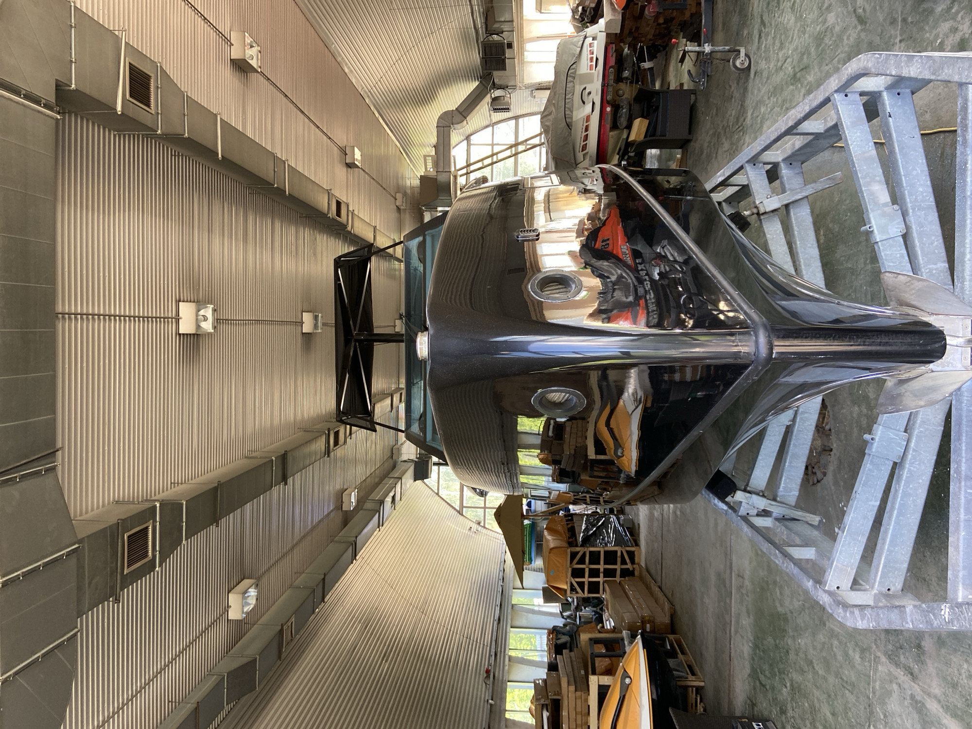 This is a forward-facing, eye-level, full shot of a black speedboat on a trailer inside a large, industrial-looking building. The boat's hull is highly reflective, showing the surrounding environment and other objects. The building has a corrugated metal roof with exposed beams and ductwork, and fluorescent lighting fixtures are visible. The boat appears to be in a workshop or storage area, with other boats, jet skis, wooden crates, and equipment visible in the background. The floor is concrete. The scene suggests a boat repair or storage facility. The location is in Votkinsk, Russia.
