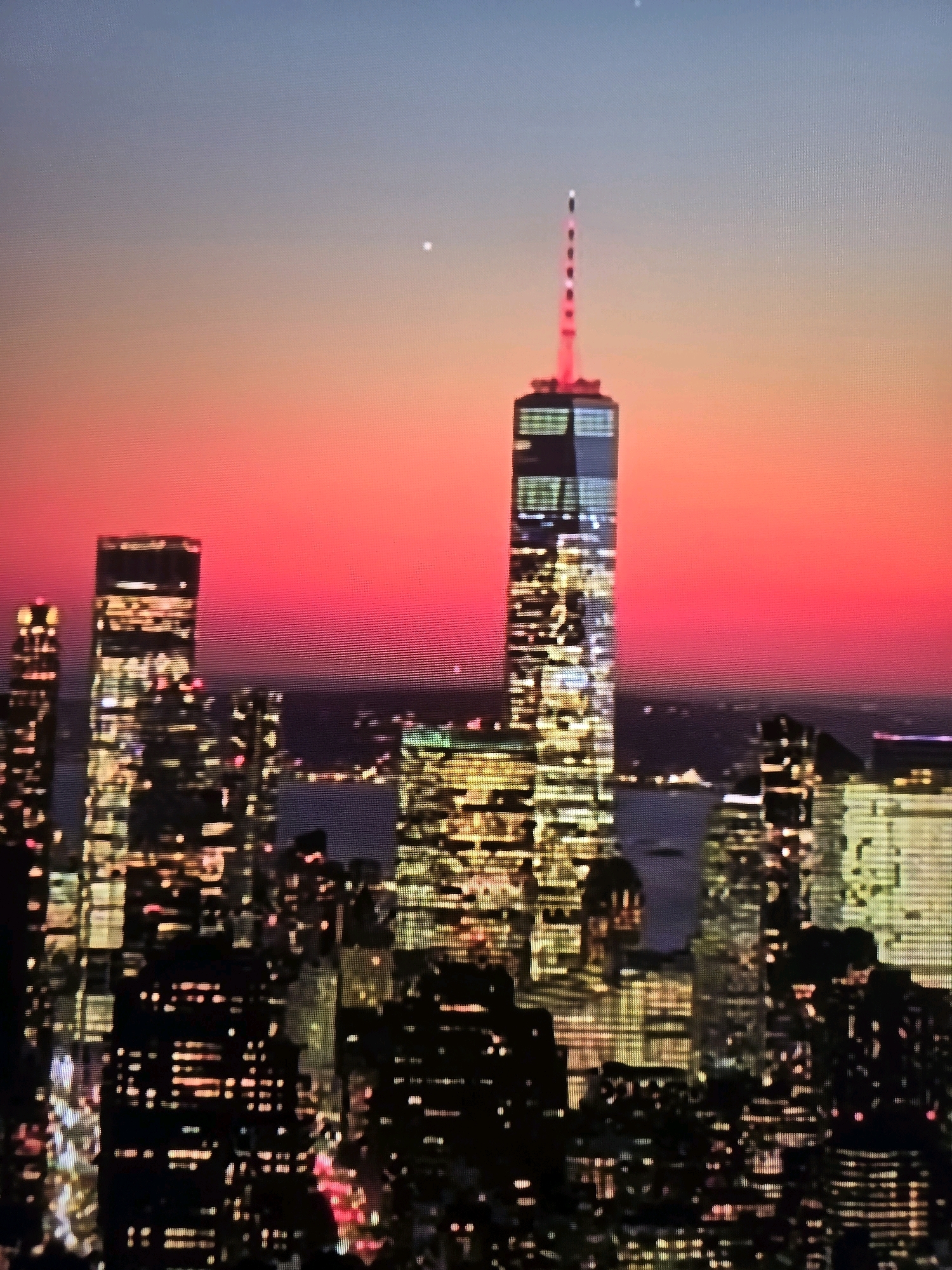 The image is a photograph of a city skyline at dusk, with a prominent skyscraper topped with a red light at its apex. The sky transitions from a deep blue at the top to a vibrant pink and orange gradient towards the horizon. Numerous other illuminated skyscrapers are visible in the foreground and midground, creating a dense urban landscape. The lights of the buildings are a mix of yellow, white, and some red. There is a hint of water in the distance, suggesting the city is on a coast or near a large body of water. The photograph appears to be taken from an elevated position, possibly another tall building or a drone. The overall impression is of a bustling metropolis at nightfall. The claim that this image is of the city of Ekibastuz, Kazakhstan is incorrect, as the architecture and style of the skyscrapers strongly resemble New York City, particularly the One World Trade Center.