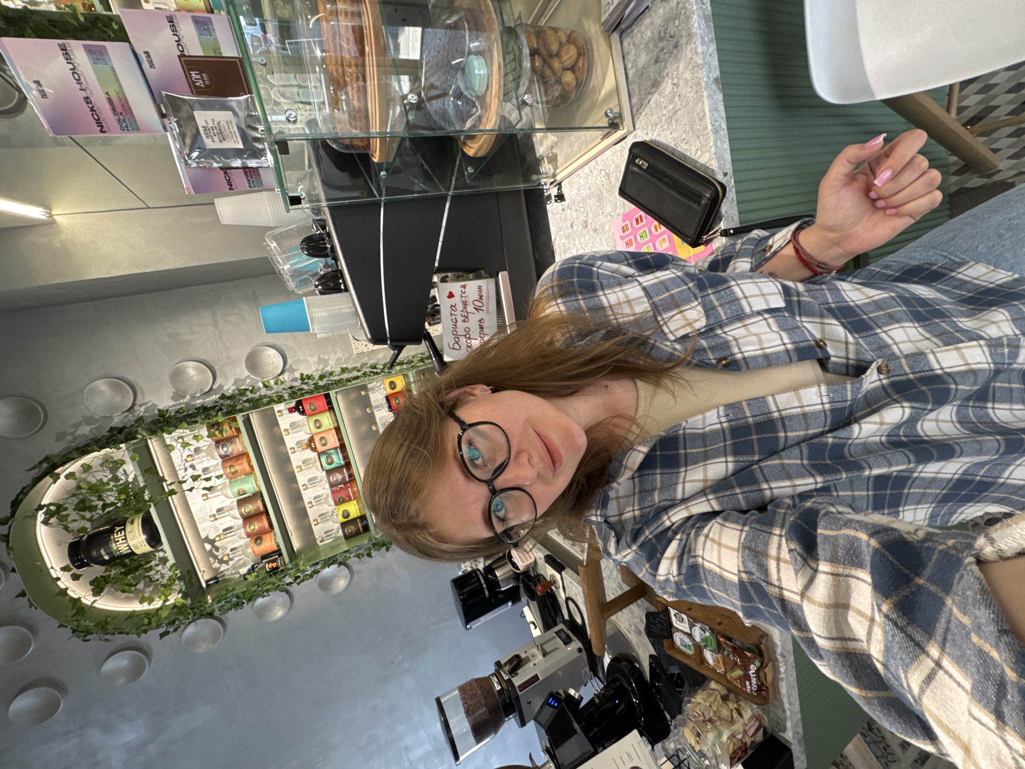 A woman with blonde hair and glasses is taking a selfie in a cafe. She is wearing a blue and white plaid shirt over a beige top. She is holding her phone in her right hand, and her left hand is resting on her chest. The cafe has a modern design with a lot of greenery. On the shelves behind her, there are bottles of various liquids and a large bottle of whey protein. In the foreground, there is a coffee machine, a display case with pastries, and a wallet on the counter. The woman is smiling and looking at the camera. The scene suggests a relaxed and friendly atmosphere. The cafe appears to be located in Perm, Russia, as indicated by the Cyrillic text visible on a sign in the background, which translates to "Barista will be back in 10 minutes."