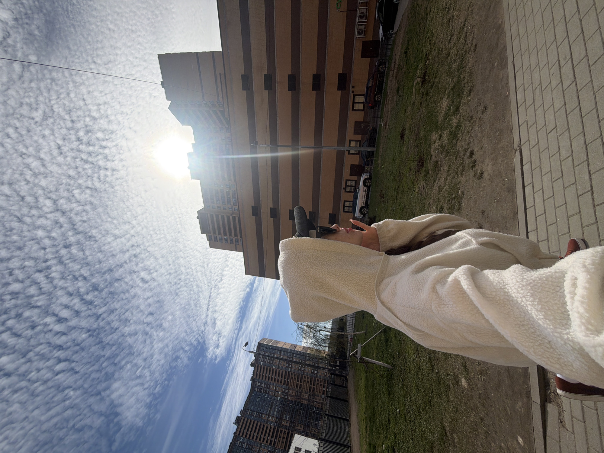 A low-angle, rotated outdoor shot features a person in a white, hooded sherpa-style jacket. The person has one hand raised making a peace sign. The background shows an urban environment with tall buildings, including a tan and brown apartment building with a grid-like window pattern. A second, darker building is also visible. The sky is filled with thin, scattered clouds, and the sun is bright. Ground cover includes green grass, pavement, and what appears to be a brick sidewalk. Several parked cars are visible near the apartment building. The person is wearing maroon sneakers and white socks. The location may be Saint Petersburg, Russia, based on the architectural style of the buildings.