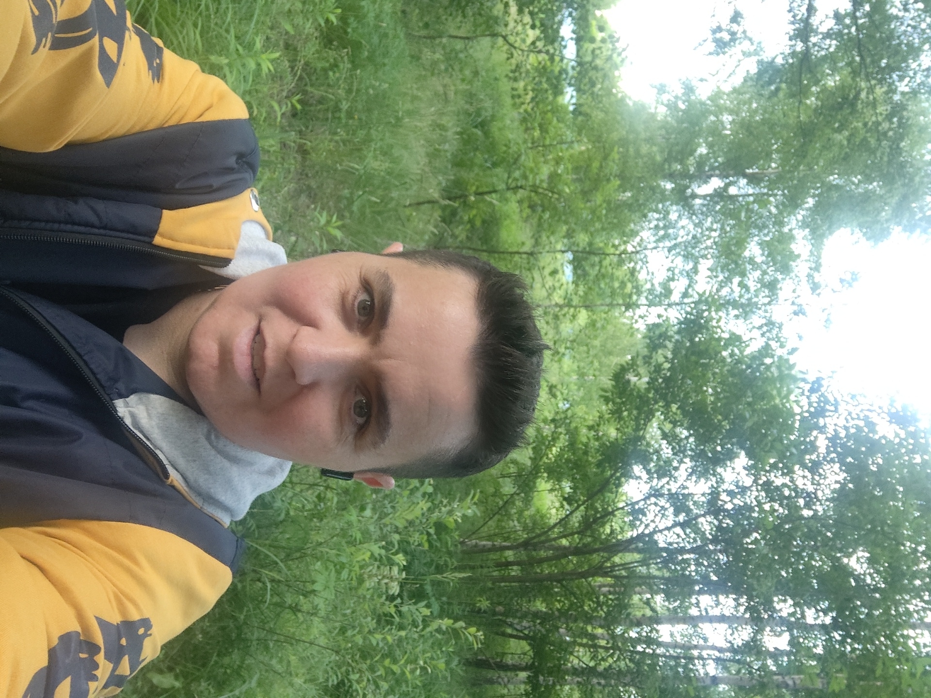 A mid-shot, front-facing selfie captures a person with dark, short hair and fair skin, wearing a yellow and dark blue jacket with a grey hood. The person is outdoors, with a blurred background of green trees and foliage, indicative of a natural environment. The setting appears to be a park or wooded area, potentially in or near the village of Tsibino, Russia. The person is looking directly at the camera with a neutral expression, and a black earbud is visible in their right ear.
