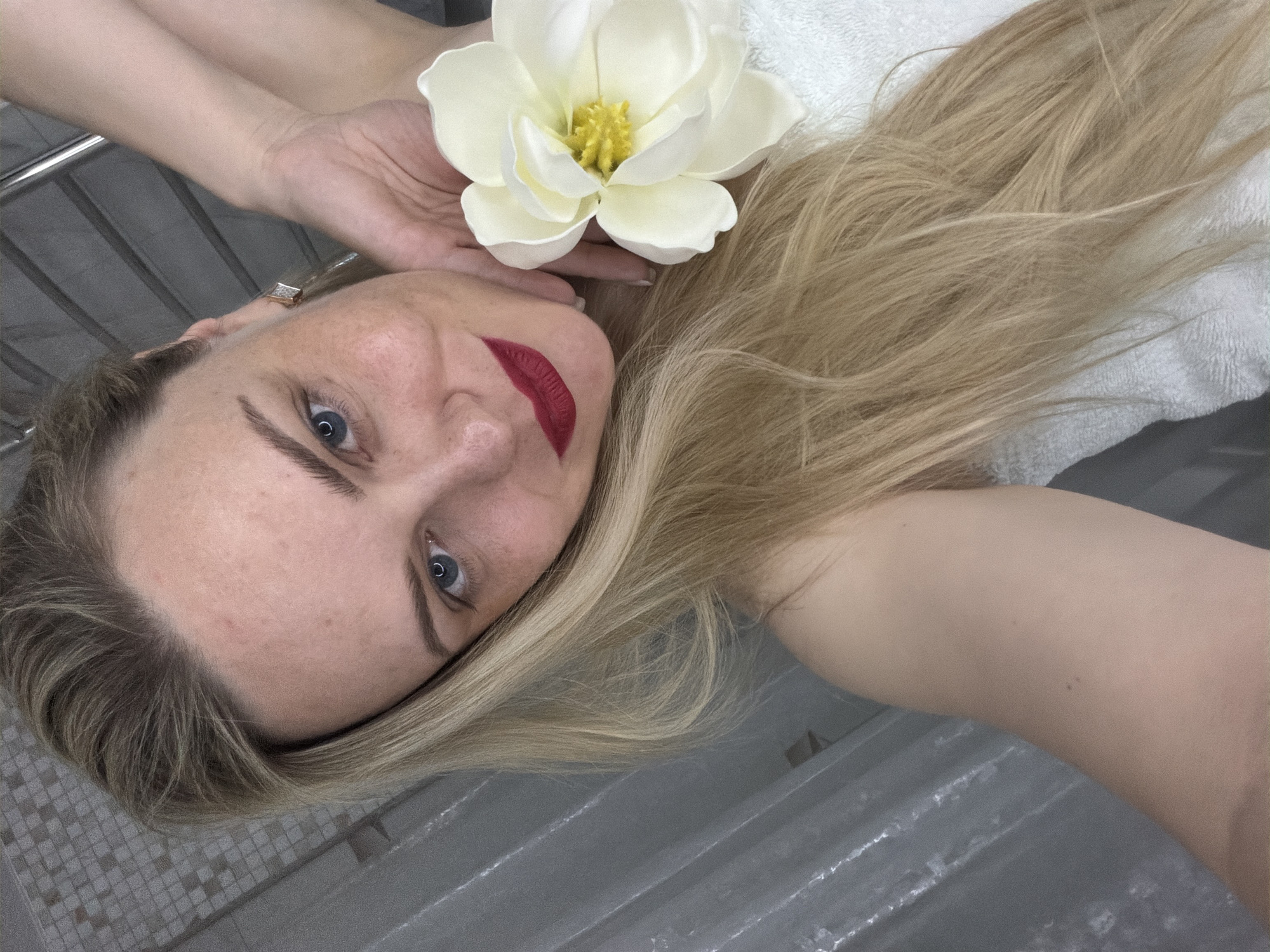 A fair-skinned woman with blonde hair is positioned in the foreground of the image, lying on a surface covered by a white towel. She has blue eyes, is wearing red lipstick, and has a white flower positioned near her face, held by one hand. The background appears to be a gray tiled surface, potentially within a bathroom setting. It is not possible to determine if the location is in the city of Votkinsk, Russia.