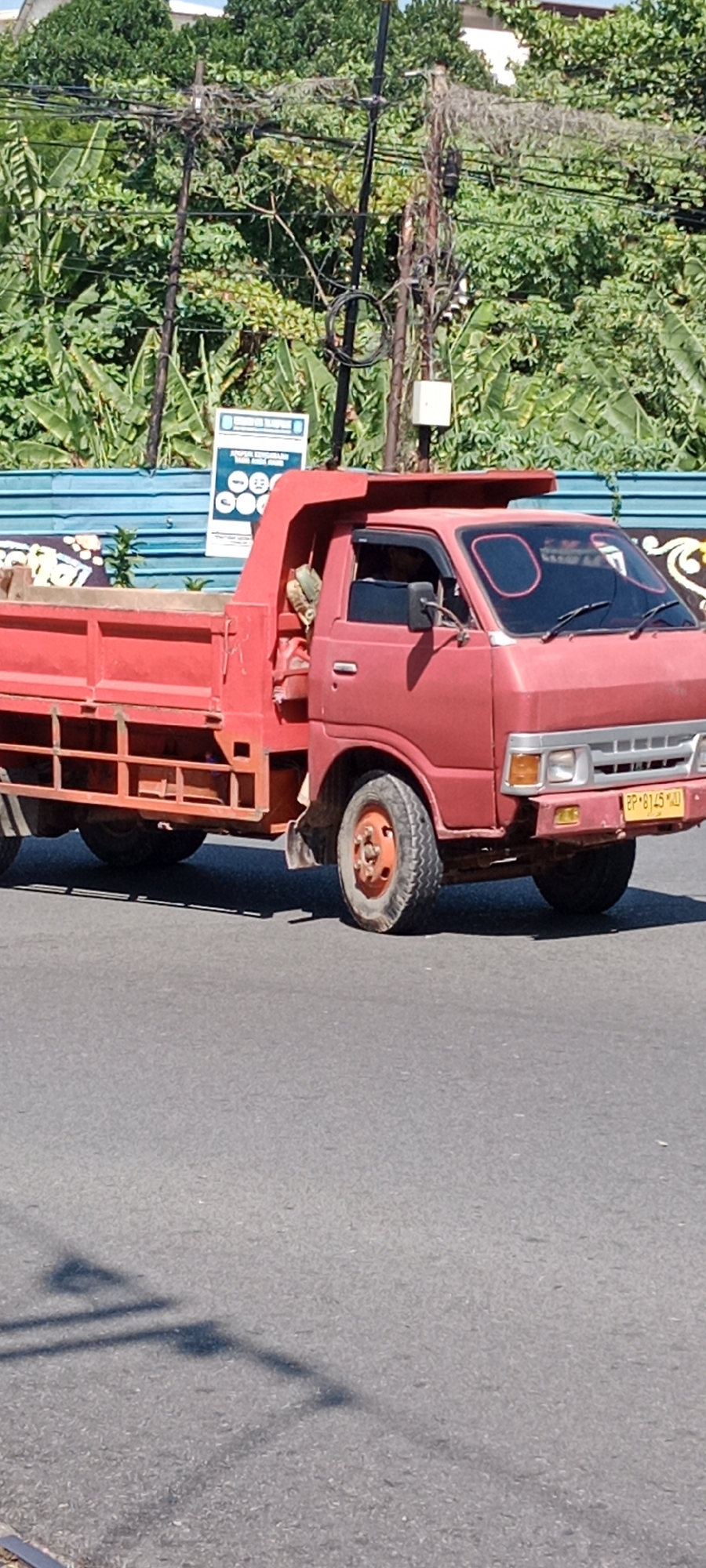 A red dump truck with a yellow license plate, "BP 8145 MU", is parked on a paved road in Tanjung Pinang, Indonesia. A person wearing a light-colored shirt is visible inside the driver's cab. The truck's bed is empty. In the background, lush green vegetation and trees are abundant. There are utility poles with numerous electrical wires crisscrossing overhead. A blue corrugated metal fence runs along the edge of the road, behind which is a white sign with a blue border. Two pink circular markings are visible on the truck's windshield. The shadow of a utility pole is cast on the road in the foreground.