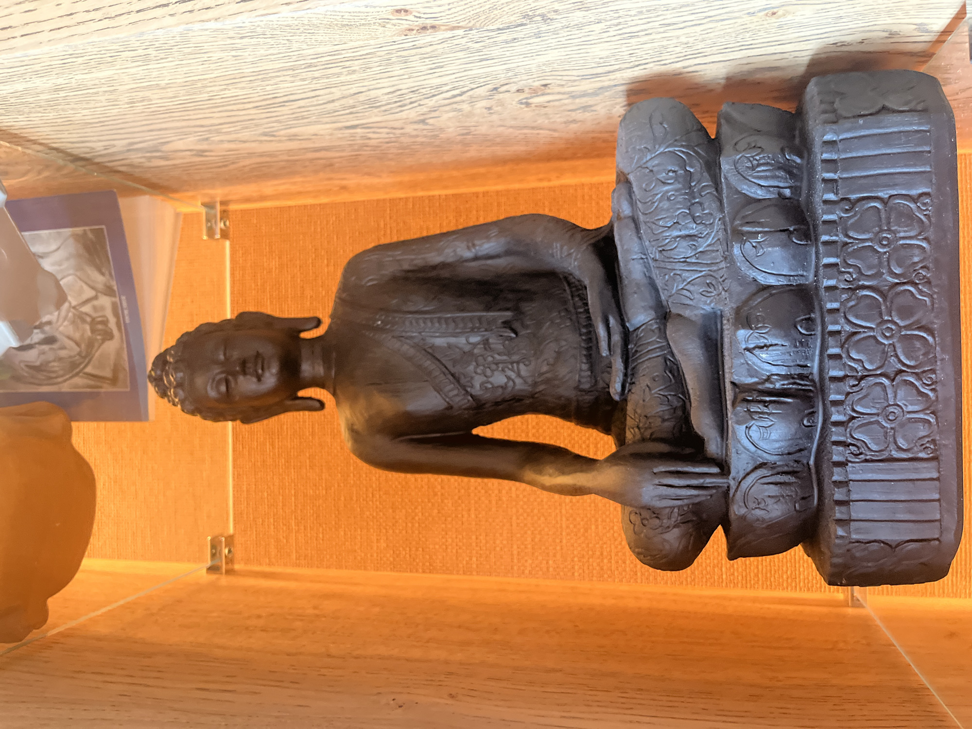 A dark-colored statue, likely depicting a Buddha or similar deity, is centrally positioned on a light wooden shelf or surface. The image appears to be taken within a display case, as evidenced by a subtle glass pane in front of the statue, supported by small metal brackets.
The statue itself is meticulously detailed, appearing to be made of a dark material such as bronze, dark wood, or resin. The figure is seated in a meditative pose, with a serene expression. Its hair is styled in tightly coiled curls. The clothing features intricate patterns, particularly visible on the chest and over the legs. The hands are placed in front of the body, possibly in a mudra, with one hand resting on the other. The base or pedestal on which the figure sits is also highly ornamented with decorative motifs, including repetitive floral patterns resembling four-petaled flowers and swirling organic designs.
Behind the statue, a textured wall with a light orange or warm brown hue serves as the background. To the left of the statue, partially visible through the glass, is a piece of paper or card with an illustration, possibly displayed in a frame or stand, and a small, light brown ceramic or pottery urn.
The scene is indoors, likely in a home or gallery setting. The lighting is warm and even, highlighting the details of the statue and the textures of the surrounding surfaces. No discernible text is visible in the image. The overall impression is one of a carefully curated display of cultural or spiritual artifacts.
