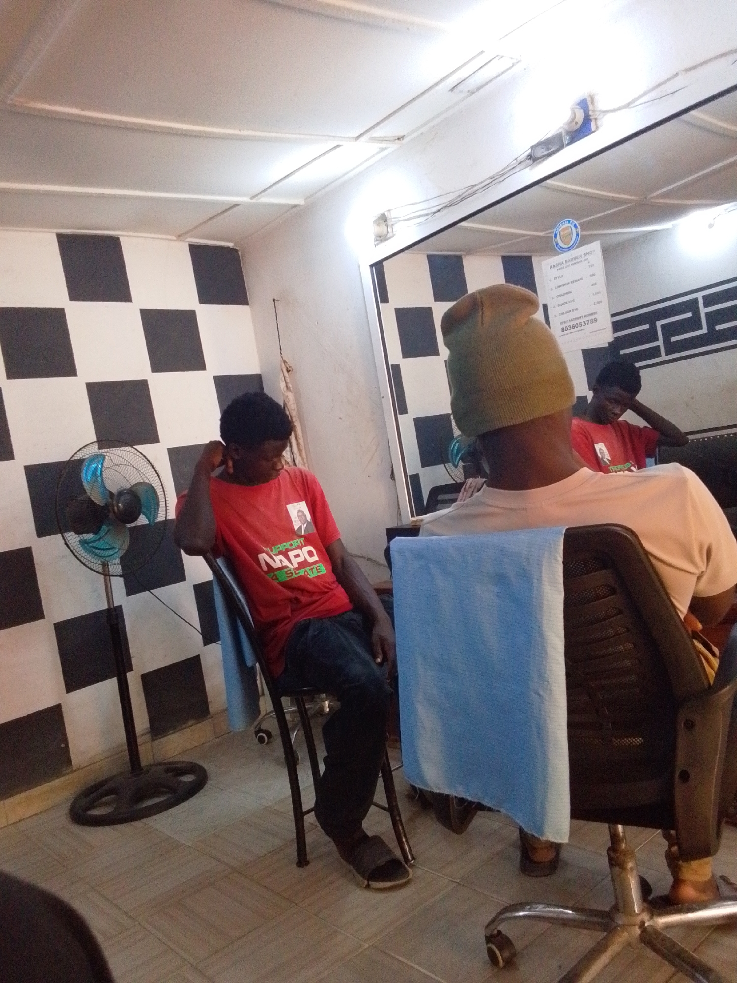 The image depicts the interior of a barber shop, likely in Jos, Nigeria, given the context. Two men are present, both appearing to be patrons. In the foreground, a man is seated in what appears to be a barber chair, with his back to the camera. He wears a light-colored t-shirt and a dark beanie, and a blue towel is draped over the back of his chair.
In the mid-ground, another man is seated in a regular chair, looking down and appearing contemplative or tired. He wears a red t-shirt that prominently displays "SUPPORT NAPO FOR SENATE" along with a picture of a man, indicating a political campaign. His reflection is clearly visible in the large mirror on the right side of the room.
The barber shop features a distinctive black and white checkered pattern on one wall. A black standing fan with blue blades is positioned in front of this wall. The floor is covered with light-colored square tiles. The ceiling is white with visible white beams and exposed wiring for light fixtures, two of which are visible.
A sign is posted on the wall, reflected in the mirror, titled "KASHA BARBER SHOP." It lists services and prices:
- STYLE: 700
- LOW/MED HAIR: 800
- CHILDREN: 1,500
- BLACK DYE: 1,500
- COLOUR DYE: 2,000
Below this, it states "PAY ACCOUNT NUMBER: 8038053789." The currency unit is not explicitly stated but is likely Nigerian Naira.
The setting suggests an ordinary moment in a local establishment, with artificial lighting illuminating the scene. No active haircutting is taking place; the men appear to be waiting or resting.