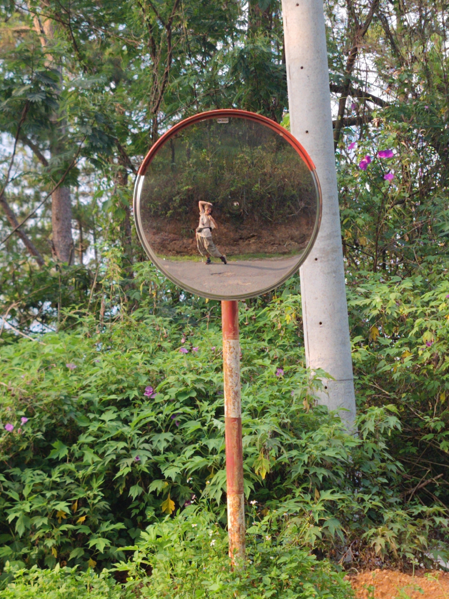 In Cam Ly - Da Lat, Vietnam, a convex mirror is positioned on a red and white pole beside a concrete utility pole, surrounded by dense foliage. The mirror reflects a person standing on a road, striking a pose with one hand on their head and another on their hip. They are wearing loose-fitting clothing. The background includes trees and vegetation consistent with the local environment.