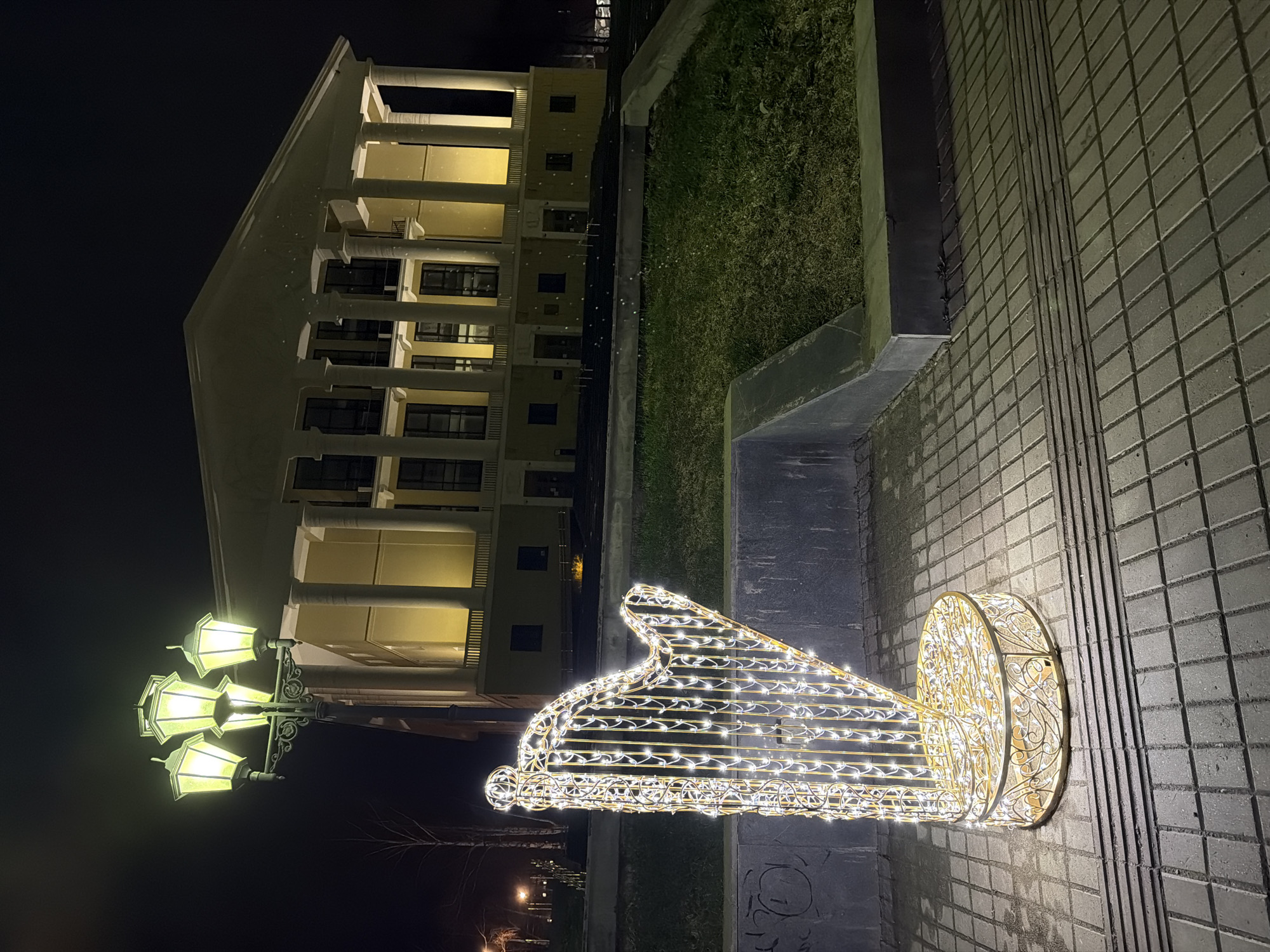 The media file presents an outdoor night scene in Votkinsk, Russia. A large, multi-story building with classical architectural elements, including prominent columns, occupies the background, illuminated from within by warm yellow light. The dark night sky above the building is dotted with numerous small white specks, possibly indicating falling snow or rain reflecting light.
In the foreground, a decorative light sculpture, intricately shaped like a grand piano, stands on a paved walkway. The sculpture is constructed from a metallic frame adorned with many small, bright white lights. To its left, an ornate streetlight with four glowing lamps emits a greenish-yellow light. The grey paved ground appears wet, reflecting the surrounding illumination. A strip of dark green grass runs parallel to the walkway, separating it from the building. No people are visible, and the scene conveys a tranquil, possibly festive, atmosphere. No text is visible in the image.