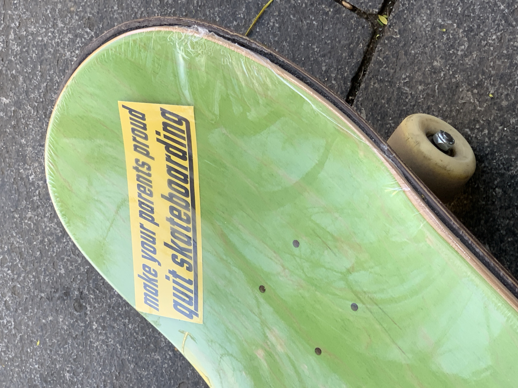 A close-up shot presents a skateboard with a light green deck, still covered in a protective plastic wrap. A yellow rectangular sticker is adhered to the deck, displaying the phrase "make your parents proud quit skateboarding" in black lettering. To the right of the deck, a single skateboard wheel is visible, appearing worn with a yellowish hue and a metal nut securing it. The background shows a section of a paved surface, possibly a sidewalk or street, with a crack running through it. The presence of the skateboard implies an urban setting, possibly used for transportation or recreation. There is no indication that the photograph was taken in Barcelona, Spain.