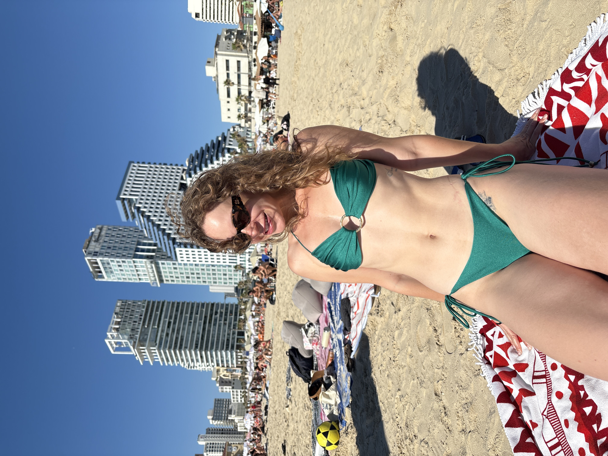 This is a portrait photograph taken on a sunny day at a crowded beach, likely in Tel Aviv, Israel. The camera is positioned at a low angle, looking upwards. The main subject is a woman with curly blonde hair, wearing sunglasses and a green bikini. She is lying on a beach towel with a red and white pattern, and her body is angled towards the camera. Her skin is tanned, and she has a faint tattoo on her side. Behind her, the beach is filled with people sunbathing, sitting on towels, and engaging in various beach activities.
In the background, a row of tall, modern white buildings with numerous windows lines the coastline, suggesting an urban beach setting. The sky is a clear, bright blue, indicating fair weather and likely midday or early afternoon. A large, dark shadow, presumably cast by the photographer, falls across the sand in the foreground, to the right of the woman. Some beach gear, including bags, shoes, and a yellow and black soccer ball, are scattered on the sand near the woman's towel. The overall atmosphere appears relaxed and leisurely, with many people enjoying the beach on a warm, sunny day.