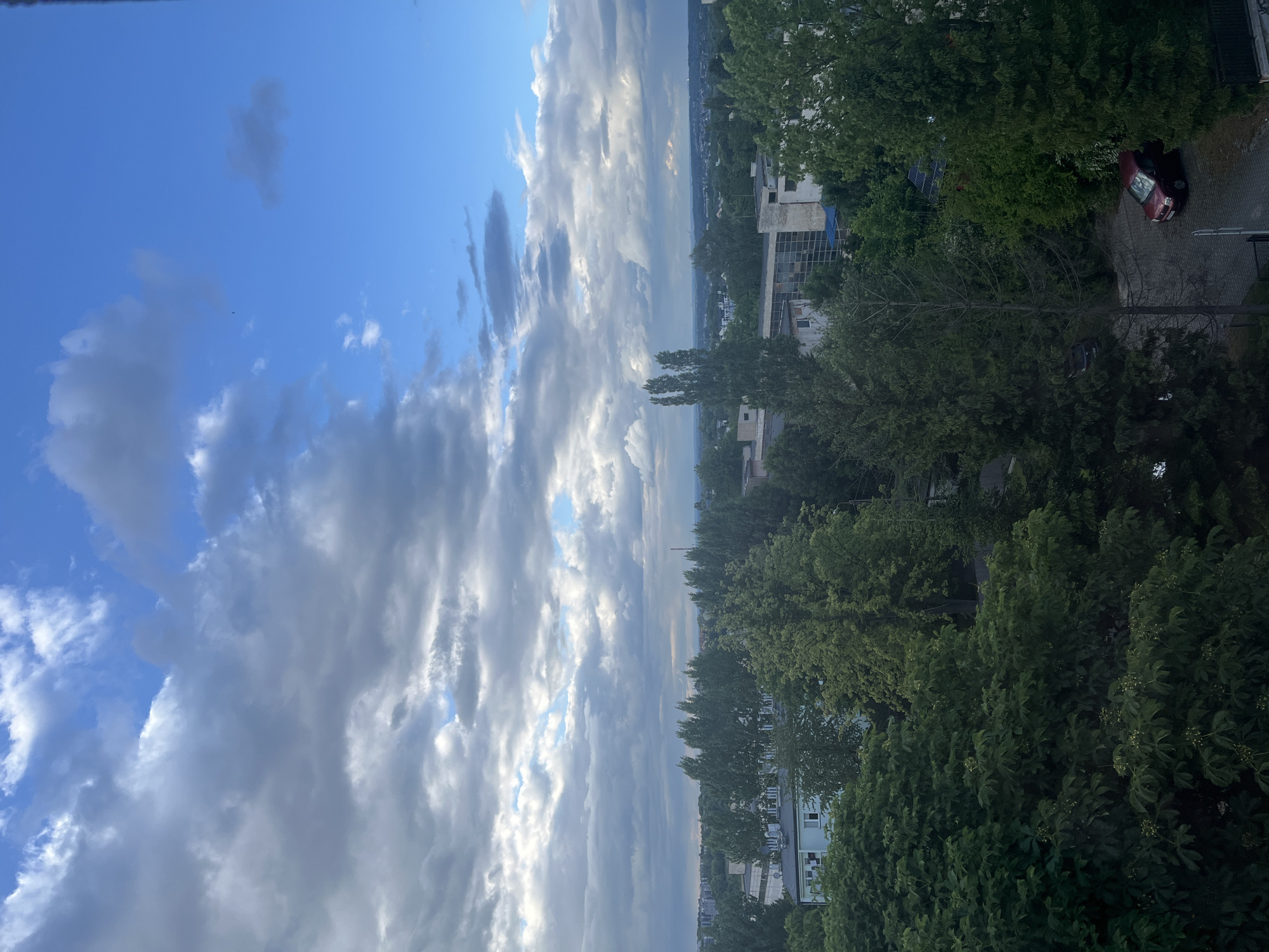 The image shows a view of a city with a cloudy sky. In the foreground, there are several large trees with dense foliage. Behind the trees, there are buildings, some of which appear to be apartment complexes or office buildings. The buildings have various architectural styles, with some featuring flat roofs and visible windows. A small red car is parked on a paved surface in the lower right portion of the image, next to a fence. The background of the image reveals a wider city landscape with more buildings and greenery extending towards the horizon, under a dramatic sky with patches of blue and a mix of dark and light clouds.