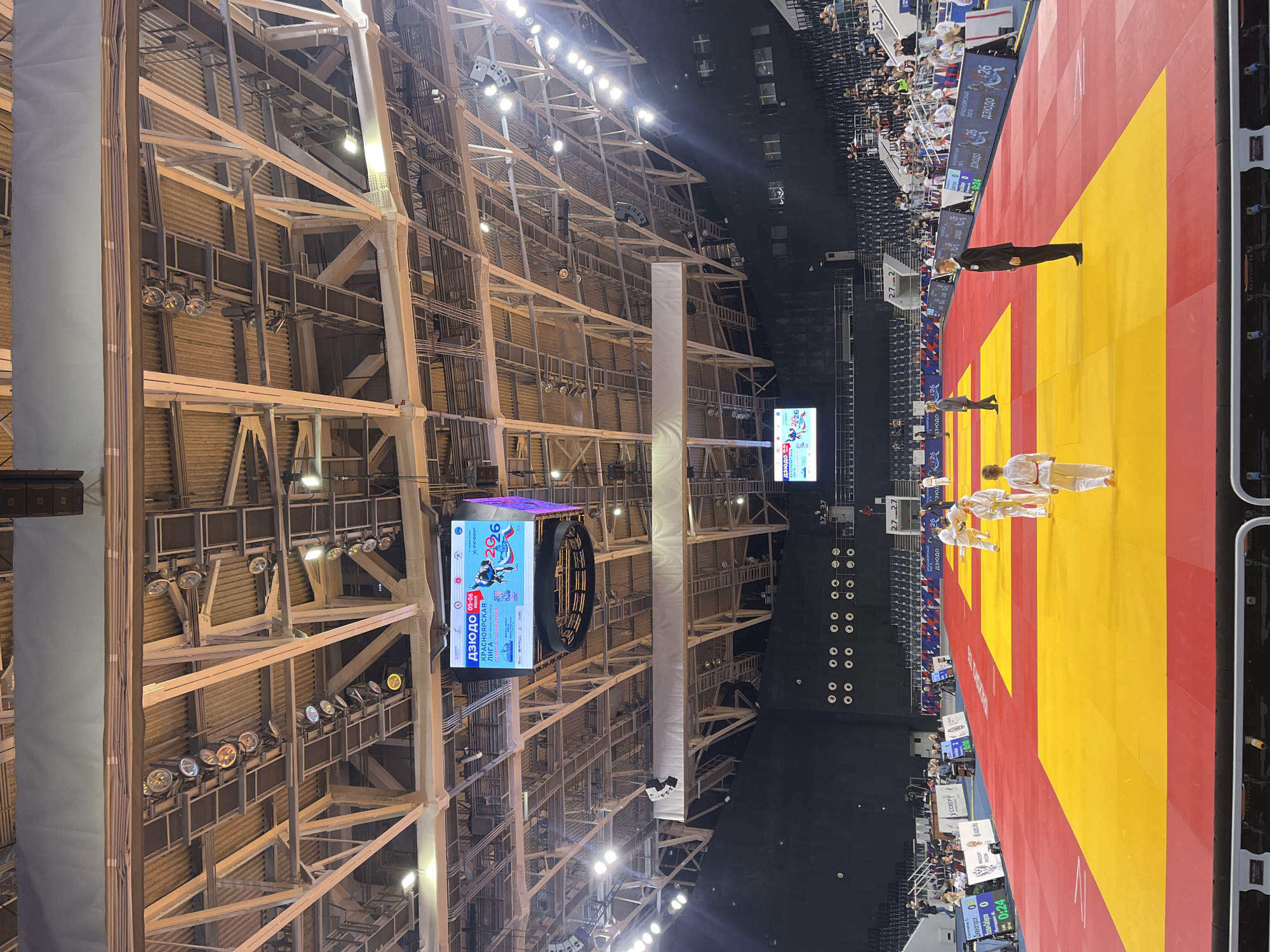 This image captures a vibrant judo competition taking place in a large, brightly lit indoor sports arena in Krasnoyarsk, Russia. The scene is dominated by a central judo mat, distinctly marked with alternating red and yellow sections.
On the mat, two pairs of judokas are visible. One pair stands facing each other, while another pair is in a bowing stance, suggesting the beginning or end of a match or a ceremonial display. All judokas are wearing traditional white and blue gis, with one pair also featuring red and white belts. Several officials in dark suits are positioned around the mat, overseeing the proceedings. Spectators can be seen in the background, seated in tiered stands.
The arena features a high ceiling with an intricate network of metal trusses and scaffolding, from which numerous spotlights illuminate the competition area. A large, circular scoreboard or display screen hangs overhead, showcasing event information. The screen displays the word "Дзюдо" (Judo), dates "05-06" (likely May 5-6), and "КРАСНОЯРСКАЯ ЛИГА" (Krasnoyarsk League), confirming the event's sport and location context. It also shows a name, "Н. БАЖЕНОВА" (N. Bazhenova), along with logos like "X СИБИРЬ" and "CDEK".
Smaller electronic displays near the mat show scores and timers, with one indicating "Дивногорск 0" and a timer at "0:24". The overall atmosphere is one of a professional and well-organized sporting event.