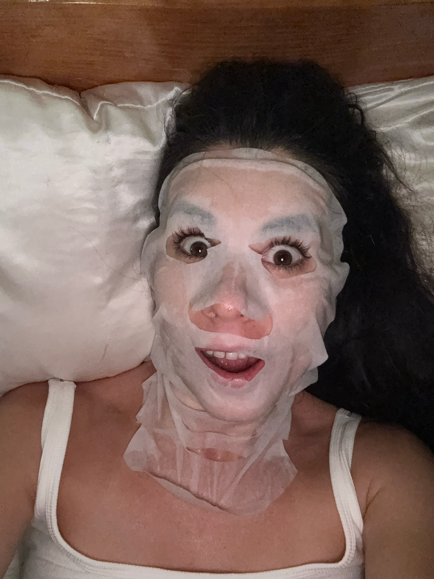 A person is lying in bed with a white sheet mask covering their face. The mask has cutouts for the eyes and mouth, and it appears to be made of a thin, white material. The person has long, dark hair that is spread out on the pillow. They are wearing a white tank top. The background is a wooden headboard and a white pillow. The image is taken from a high angle, looking down at the person.