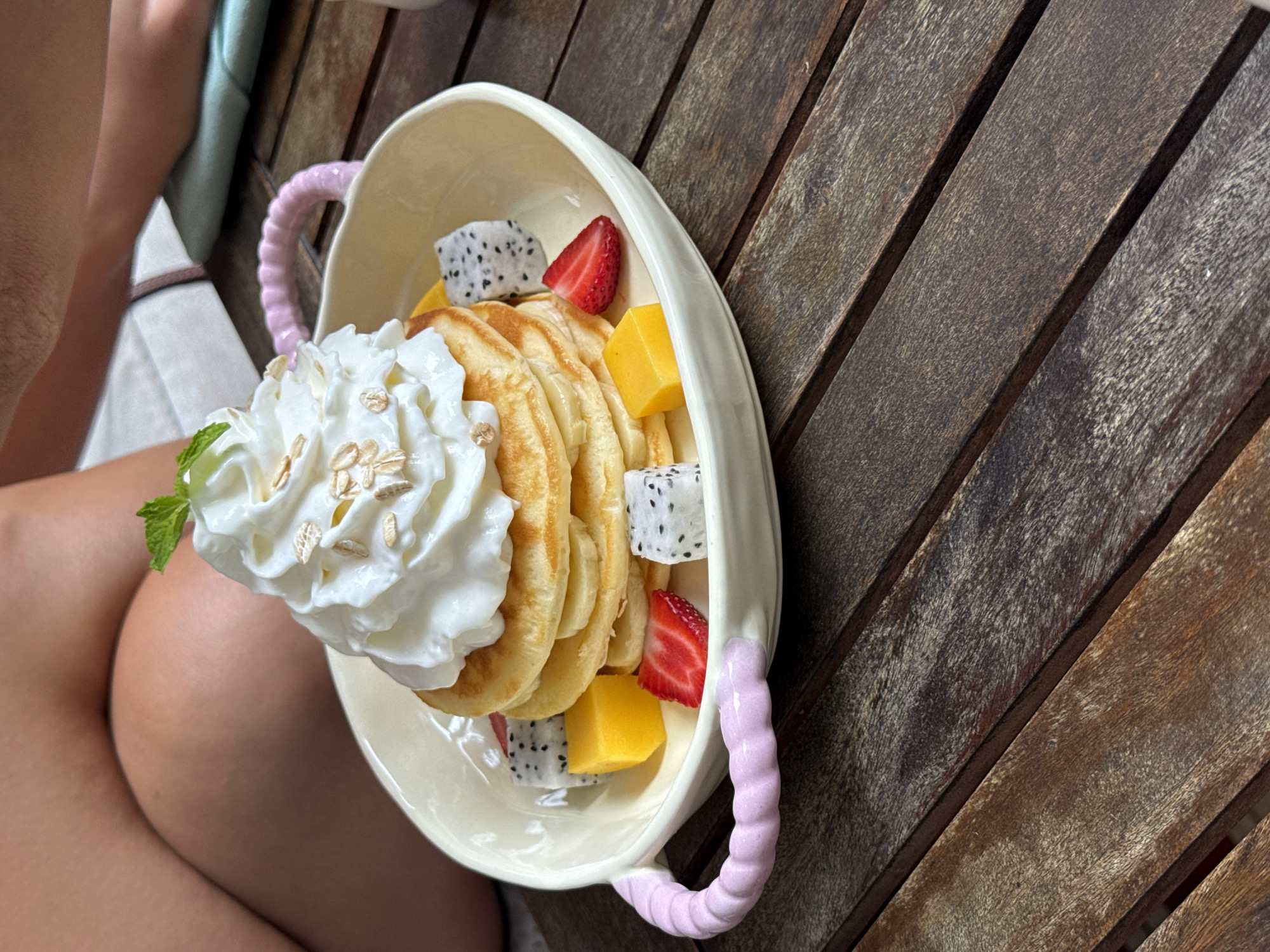 The image shows a plate of pancakes with whipped cream and fruit on a wooden table. The fruit includes strawberries, mango, and dragon fruit. The pancakes are topped with oats and a mint sprig. The plate is oval-shaped with pink handles. The table appears to be outdoors, constructed of wooden slats. The location is Đà Nẵng, Vietnam.