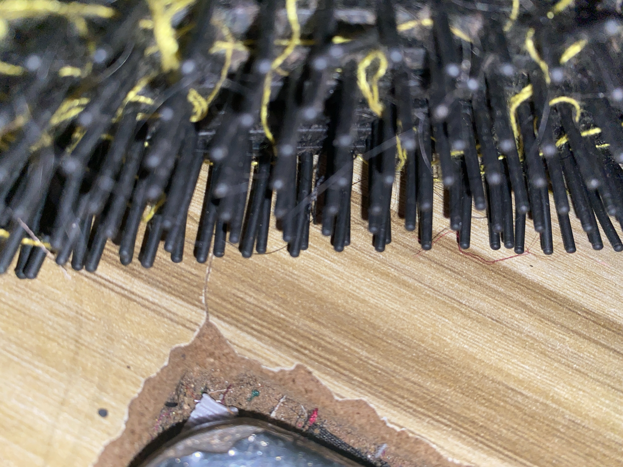 The image is a close-up, angled shot of a damaged wooden surface with a hairbrush positioned on it. The wooden surface has a light brown, grainy texture with visible parallel lines. In the upper left corner, a portion of a shiny, possibly glass or plastic object with glitter embedded in it is visible. Adjacent to this object is a torn edge of what appears to be paper or cardboard, with small debris scattered on it, including specks of color like red and green.
The hairbrush dominates the right side of the image and occupies the majority of the frame. It has black bristles, and interspersed among them are strands of bright yellow material, possibly lint or colored threads. Some of the bristles have small white tips. The bristles are densely packed and appear slightly worn or dirty.
The scene appears to be indoors, given the nature of the objects. There are no people or animals visible in the image. The lighting is somewhat diffused, creating soft shadows. The overall impression is one of everyday objects in a state of disarray, possibly indicating a discarded or neglected area. The specific location in Jauro Yinu, Nigeria cannot be determined from the visual information provided in the image.