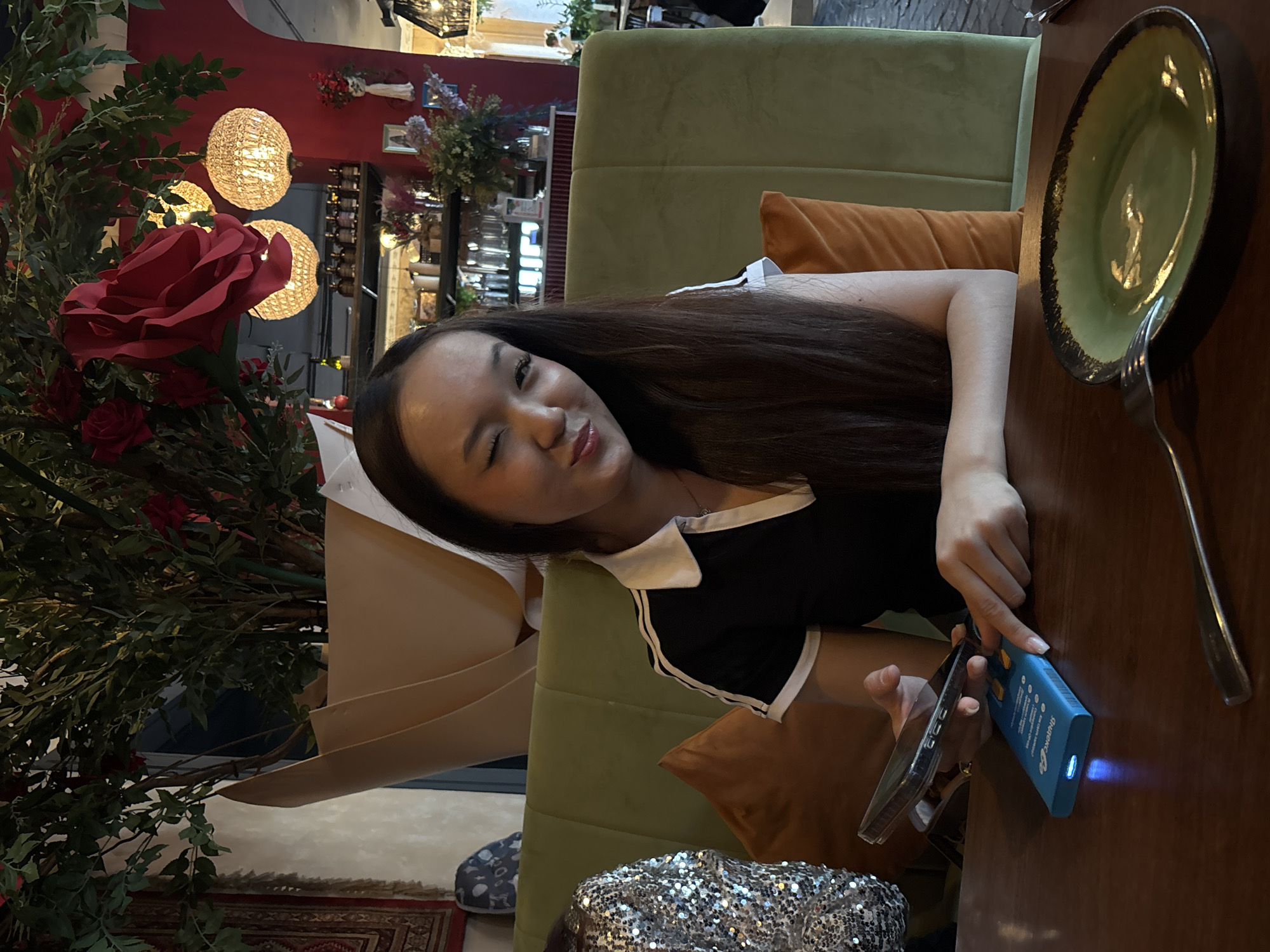 This image depicts an indoor scene within an establishment located in the city of Saint Petersburg, Russia, likely a restaurant or cafe. A young woman of East Asian or Southeast Asian descent is seated at a dark wooden table, directly facing the camera with a wink of her left eye and a slight smile. She has long, dark brown hair and is wearing a black polo shirt with a white collar and white trim, including two white stripes on the sleeves.
Her left hand holds a black smartphone encased in a clear cover. Her right hand rests on a rectangular blue device with yellow buttons and a glowing blue light on one end, possibly a power bank or payment terminal, which has "Sharge..." text partially visible. On the table to the right, an empty oval plate with a green center and a dark rim is present, alongside a silver fork and knife.
The woman is seated on an olive-green upholstered booth seat. To her immediate left, part of another person's arm or shoulder, clad in a silver sequined garment, is visible. The background features extensive decor. Behind the woman, a large potted plant with abundant green foliage and several prominent artificial red roses dominates the space. The pot is wrapped in light brown, textured paper. To the right of the plant, a deep red wall is visible, adorned with several spherical, ornate chandeliers emitting warm light. Further back, a bar or counter area with bottles, glassware, and floral arrangements, including purple and pink flowers, is visible. On the far left, a patterned tapestry or rug hangs on a lighter-colored wall, below which a grey and white decorative object is mounted. The overall lighting is warm and ambient.