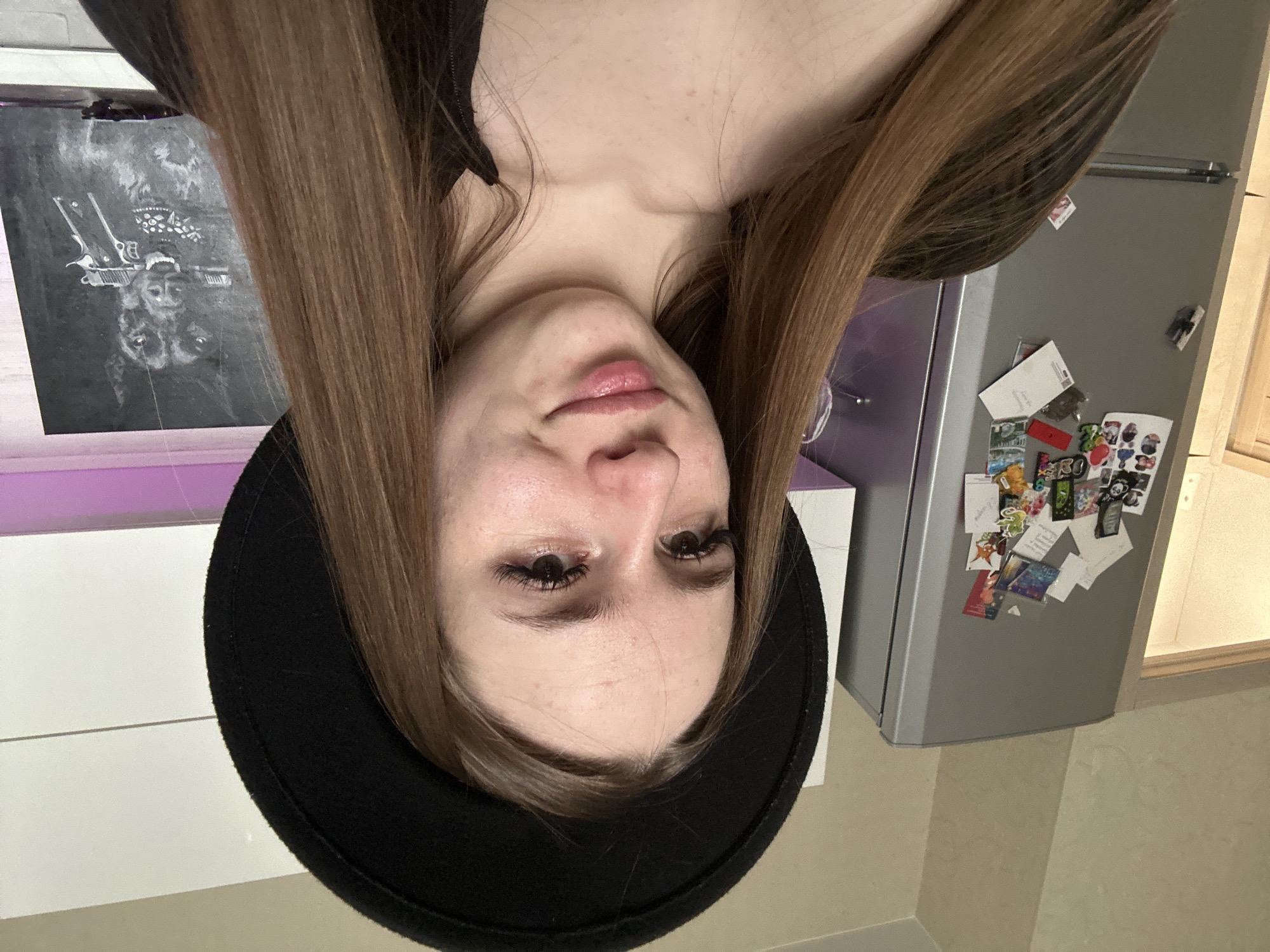 A woman with long brown hair and a black hat is centrally framed, face-up, in a rotated indoor shot. Behind her is a silver refrigerator covered with magnets and papers. To the left, a black and white artwork hangs above a white surface with a purple-toned strip. The floor is a light neutral color.