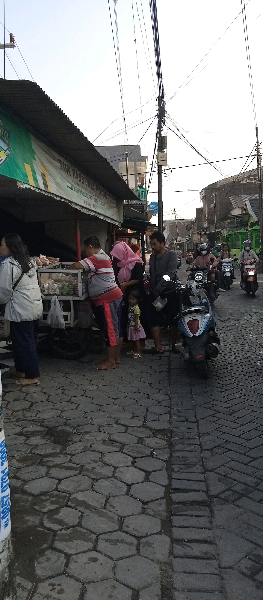 In Wadungasri, Indonesia, an outdoor scene unfolds with a street market. The foreground displays a cobbled street, partly wet, with hexagonal paving stones on the left and rectangular ones on the right. A white pole with blue lettering is visible on the far left.
A vendor's stall is prominent, featuring a corrugated metal roof and a banner advertising "THK PASS JAYA MOTOR" and "JUAL BELI MOBIL BEKAS CASH KREDIT" (Buy and sell used cars cash credit). Underneath the banner, a display case holds food items.
Several people are gathered around the stall. A woman in a white jacket stands on the left. A man in a red and white striped shirt is serving or arranging items from the display case. Next to him, a woman in a pink headscarf is speaking with him. A young girl in a yellow shirt and pink skirt stands between them, looking towards the stall. On the right, a man in a dark grey shirt stands behind the woman in the pink headscarf.
Further down the street, a procession of motorbikes moves past. One motorbike is stopped in the middle, a blue Honda scooter. Other motorbikes with riders, some wearing helmets, are visible further down the road, indicating traffic. The background shows buildings and utility poles with numerous wires crisscrossing overhead, typical of an urban Indonesian environment. The overall impression is a bustling street scene with commercial activity.