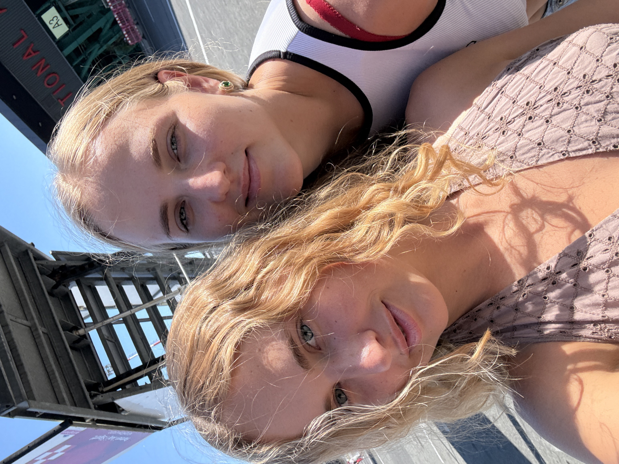 Two young women with blonde hair pose for a selfie in front of a large building with red lettering that reads "TIONAL". The building appears to be part of an international venue, possibly a stadium or airport. The woman in the foreground has curly blonde hair and is wearing a light brown dress. The woman in the background has straight blonde hair and is wearing a white tank top with a red strap visible. Both women are smiling. The scene is outdoors with a clear blue sky. The location is identified as Kruja, Albania.