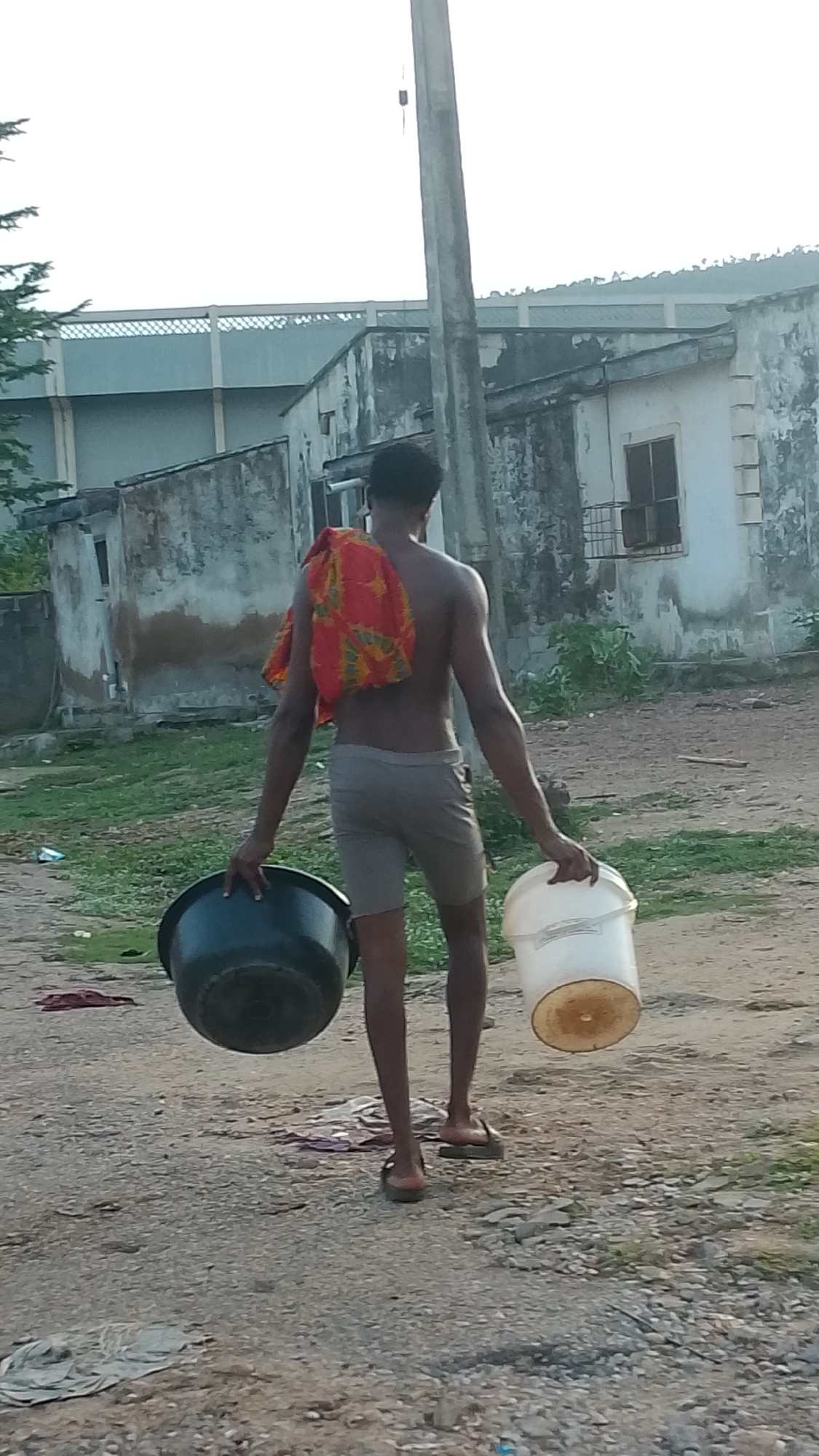 A young man is depicted walking away from the camera in a semi-urban or rural setting, likely Lokoja, Nigeria. He is shirtless, wearing light-colored shorts and dark flip-flops. A vibrant, red and orange patterned cloth is draped over his left shoulder. He carries a large, dark basin in his left hand and a white bucket with a discolored base in his right hand, suggesting he is performing a chore such as fetching water or doing laundry.
The ground is an uneven dirt or gravel path with patches of sparse vegetation. In the background, several single-story buildings with weathered, peeling walls and simple window frames are visible, hinting at a modest living environment. Further back, a larger, possibly industrial or commercial structure with a grid-like facade can be seen. A tall, grey utility pole stands prominently on the right side of the frame.
The lighting is soft and diffused, characteristic of either early morning or late afternoon, with a hazy, bright sky overhead. The overall scene conveys a sense of everyday life and routine activity in a community where infrastructure may be basic. There is no discernible text within the image.