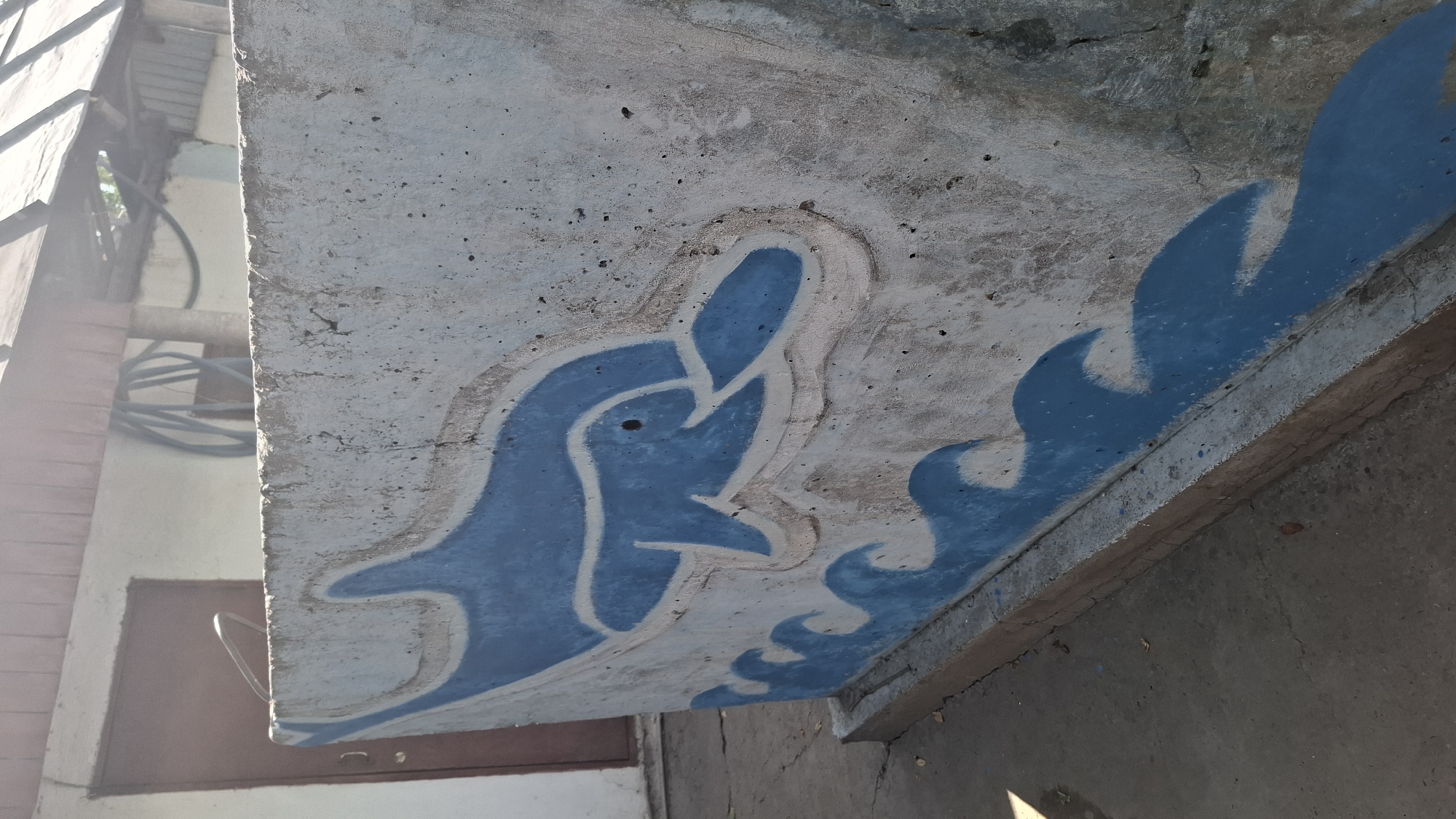 A concrete wall with a blue and white mural of a dolphin jumping over waves is depicted in this image. The wall is weathered and shows signs of age. In the background, a brown door and a portion of a building are visible. The ground in front of the wall is concrete and appears cracked. The scene is outdoors, under a sunny sky. The location is Шұбарсу, Kazakhstan.