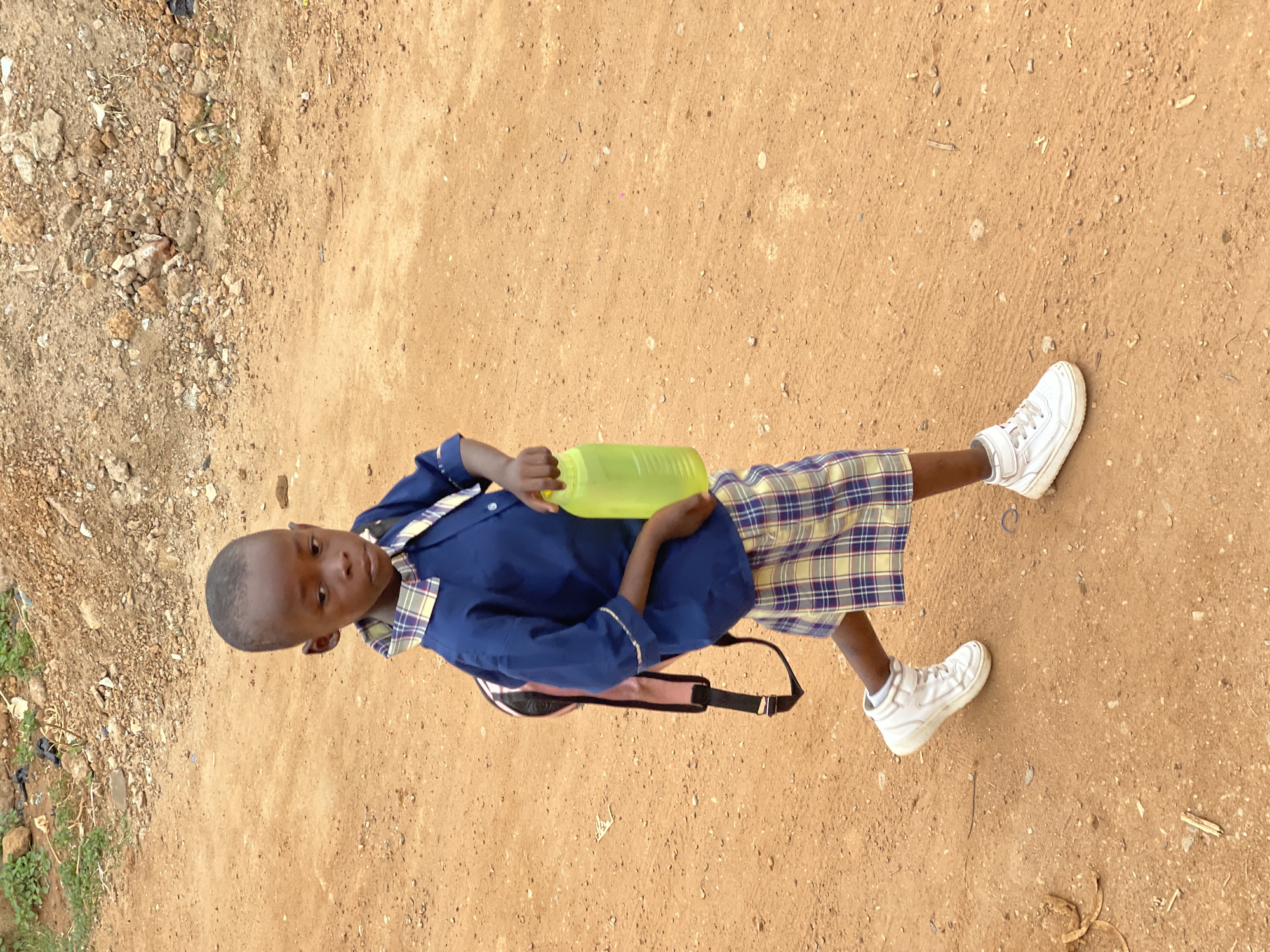 A young Black boy stands on a sandy, dirt-covered area near Kuchingoro, Nigeria. He is wearing a blue school uniform with plaid shorts and white sneakers. He is also carrying a pink backpack and holding a yellow plastic bottle. The ground is dry and dusty, with scattered rocks and debris in the background.