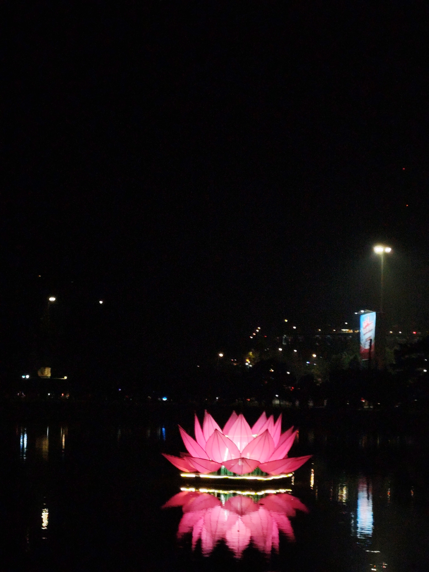 A nighttime scene depicts a large, illuminated pink lotus flower floating on a body of water. The reflection of the flower is visible on the water's surface. In the background, there are city lights, a tall streetlight, and a billboard. The scene is in the city of Da Lat, Vietnam.