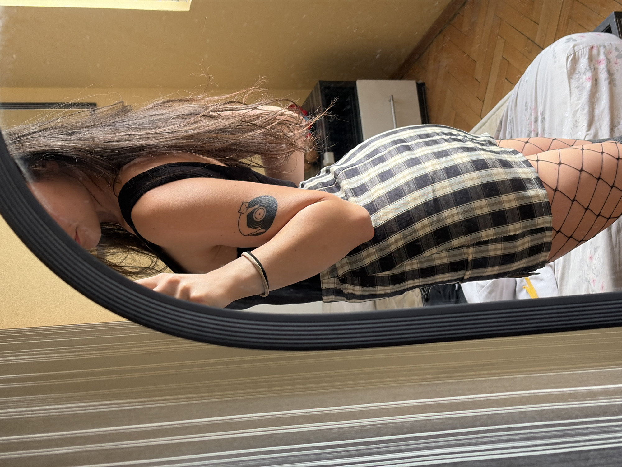 A woman is seen from the side, reflected in a mirror, in an indoor environment. She is wearing a black tank top, a plaid skirt, and fishnet stockings. A tattoo of a record with a cat on it is visible on her upper arm, and she is wearing a bracelet. The background includes a beige wall, a dresser with drawers, and a bed with a floral-patterned comforter. The floor is made of wood planks.