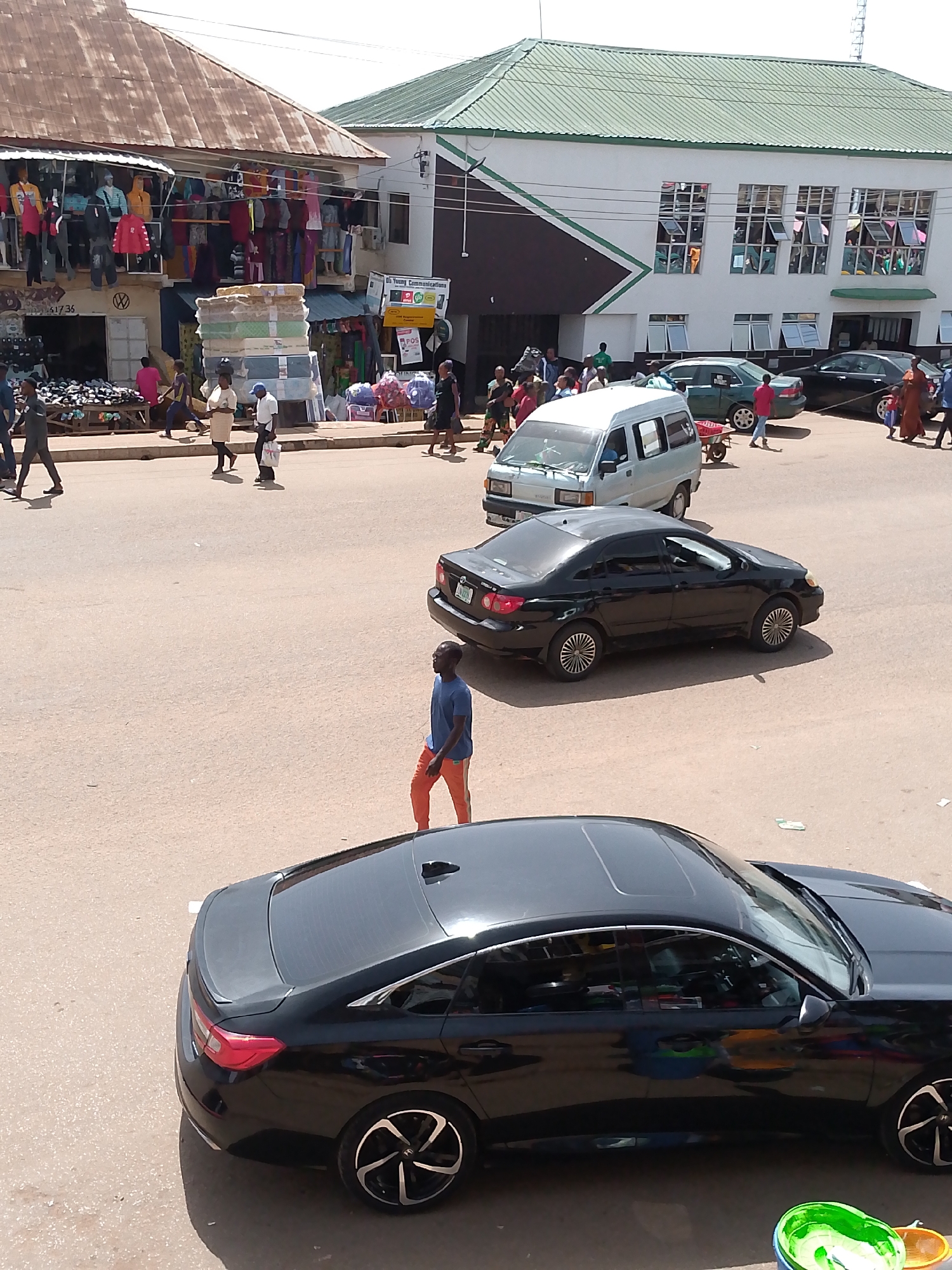 The image depicts a bustling street scene in Bukuru, Nigeria, under a bright, sunny sky, likely mid-morning or afternoon. The foreground is dominated by the rear and side of a sleek, black sedan with tinted windows and stylish alloy wheels. Beyond this car, a man in a blue t-shirt and orange trousers walks across the road, his shadow cast on the pavement.
Further back, the street is filled with a variety of vehicles, including a white minivan with a trailer attached and several cars, one of which is a black sedan. A green car is also visible. People are actively engaged in various activities: some are walking on the sidewalk, while others appear to be browsing or working at market stalls.
On the left side of the image, a row of market stalls is set up under a weathered, corrugated iron roof. These stalls display a colorful array of clothing on hangers, as well as stacks of mattresses. Signs are visible on some of the stalls, including one that reads "Dr. Young Communications" and another with "POS" written on it. The buildings in the background have green roofs, and the overall impression is one of vibrant commerce and daily life unfolding in an open-air market environment. The scene conveys a sense of energy and activity typical of a busy marketplace.