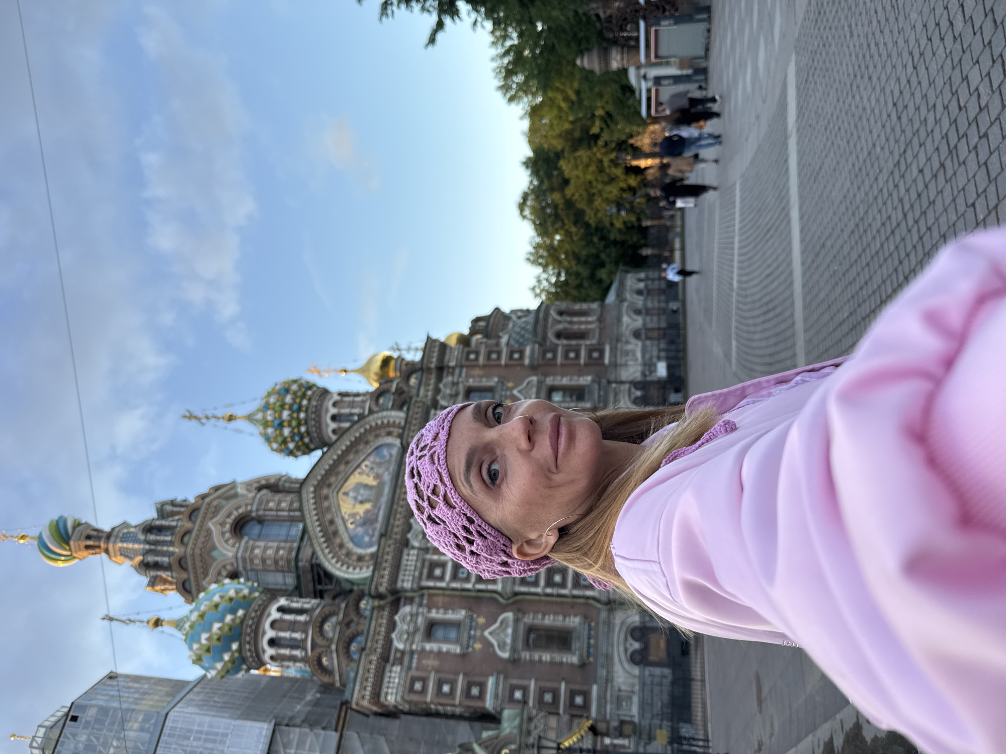 A woman in a pink jacket and a pink crochet headscarf is standing in front of the Church of the Savior on Spilled Blood in Saint Petersburg, Russia. The church is a large, ornate building with multiple domes, some of which are decorated with blue and green patterns. The sky is blue with some scattered clouds. In the background, other people are walking in a plaza paved with cobblestones. The overall lighting suggests it is either early morning or late afternoon.