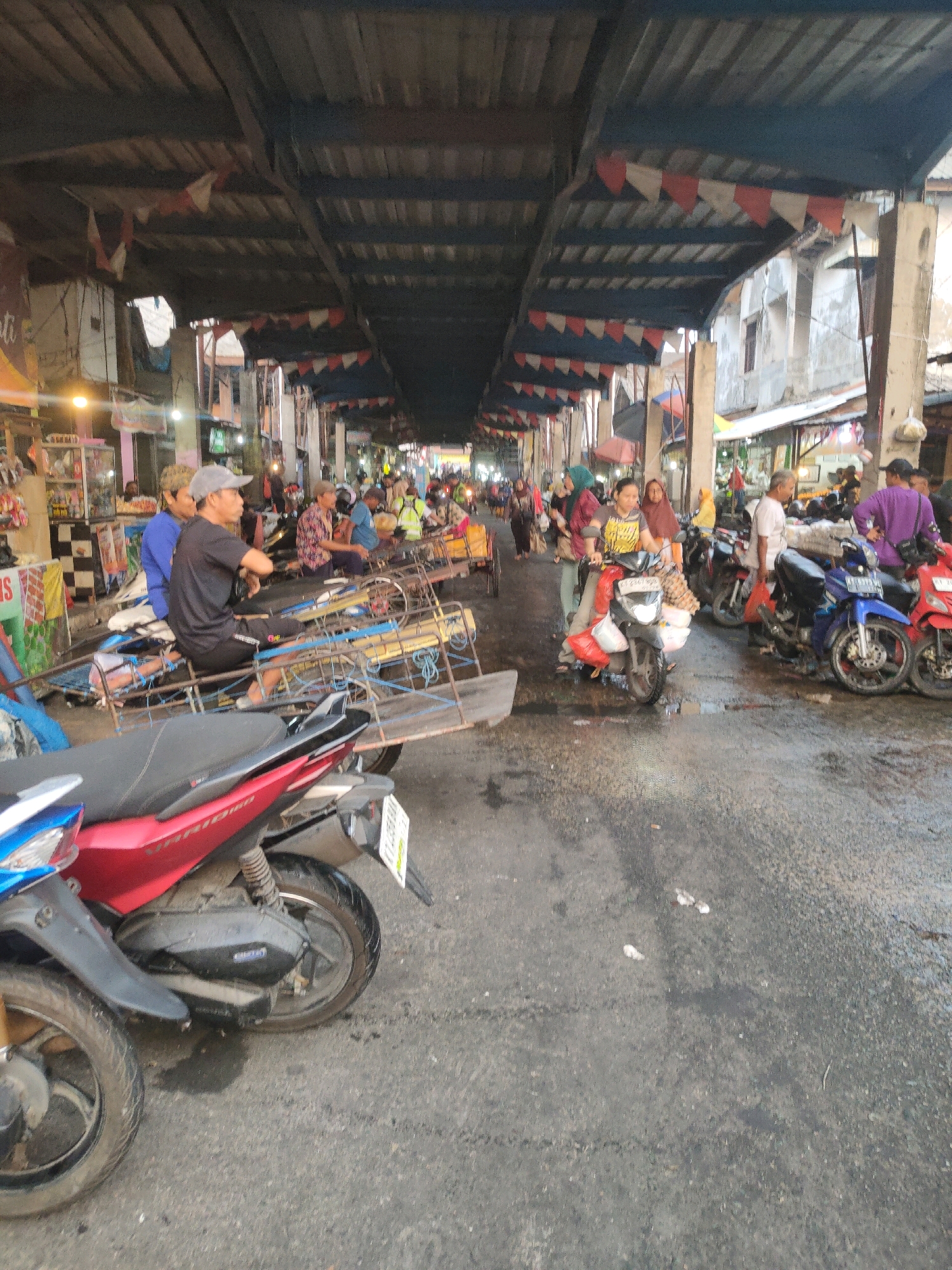 This image captures a bustling street market scene in Samarinda, Indonesia. The market appears to be located under a covered structure with a corrugated metal roof supported by pillars. Red and white triangular flags are strung along the roofline, suggesting a festive atmosphere or a specific event. The ground is paved and wet, with some puddles visible, indicating recent rain or a cleaning.
Several people are present, with a focus on those interacting with motorcycles and carts. In the foreground, two motorcycles are parked, one red and one partially visible blue, with the red one being more prominent. A man wearing a black t-shirt and shorts, with a gray cap, is seated on a motorcycle. Another man in a blue shirt is seated behind him. They appear to be waiting or resting.
Further into the market, more people are seen. A woman on a scooter, wearing a yellow and black shirt, is surrounded by bags and appears to be carrying goods, possibly groceries or market produce. Several other individuals are walking and standing among the stalls. Some are pushing carts or riding motorcycles laden with items. The stalls themselves are lined up along the street, displaying various goods, although they are not clearly discernible in detail. The overall impression is one of a busy commercial area where goods are transported and sold, with motorcycles serving as a primary mode of transportation and potentially for delivery. The architecture of the market suggests a traditional or semi-permanent setup, common in many Indonesian cities.