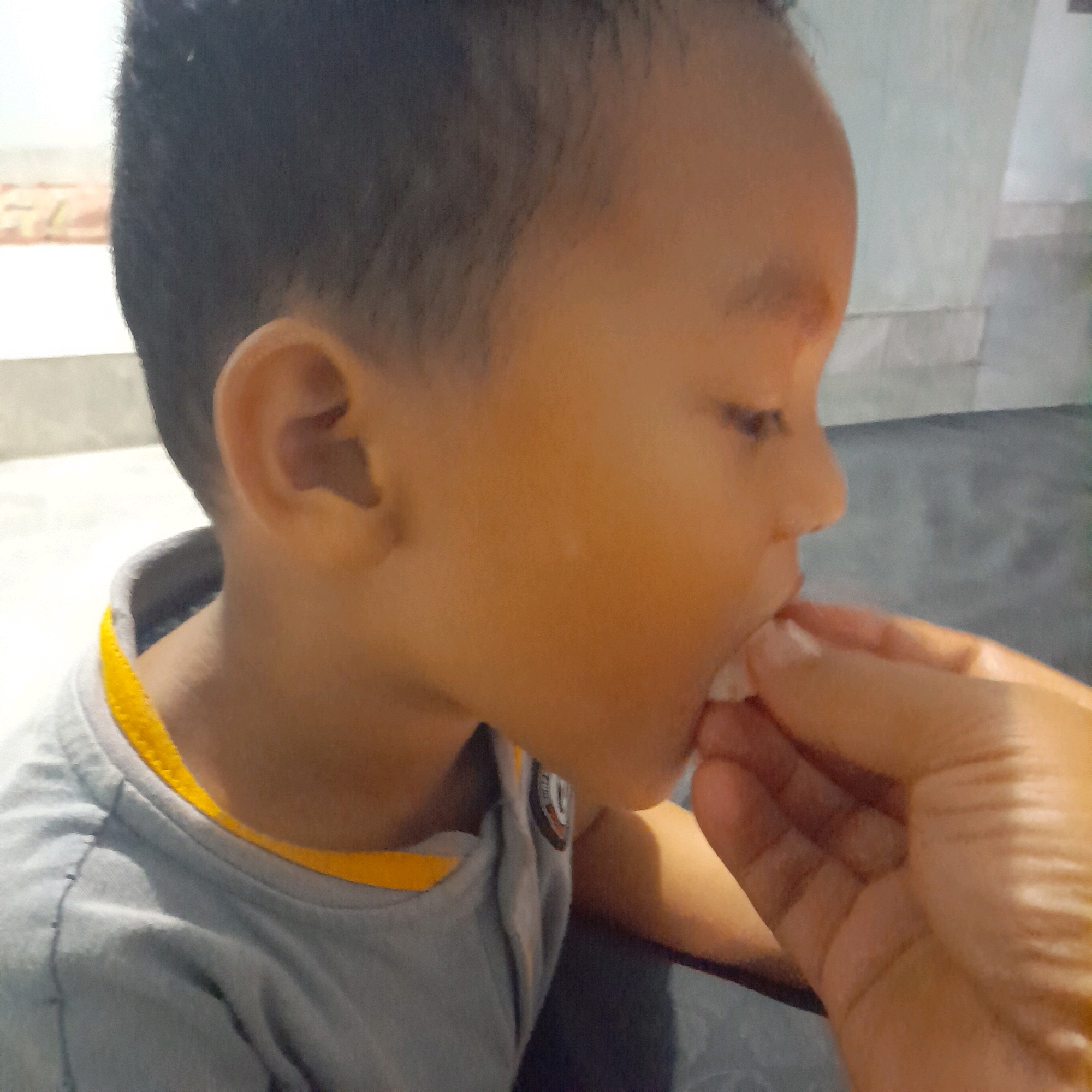 The image is a close-up shot of a young child, likely a boy, being fed something small and white by an adult's hand. The child's face is in profile, turned towards the right side of the frame. His skin tone is light brown, and he has short, dark hair. He is wearing a gray t-shirt with a bright yellow trim around the collar. The adult's hand is partially visible, with fingers extended to offer the food to the child's open mouth. The background is blurred, suggesting an indoor setting with light-colored walls and a darker surface on the right. The lighting appears to be natural, possibly from a window, casting a warm glow on the child's face. There is no discernible text in the image. The focus is on the interaction between the child and the person feeding him, capturing a moment of nourishment or care.
