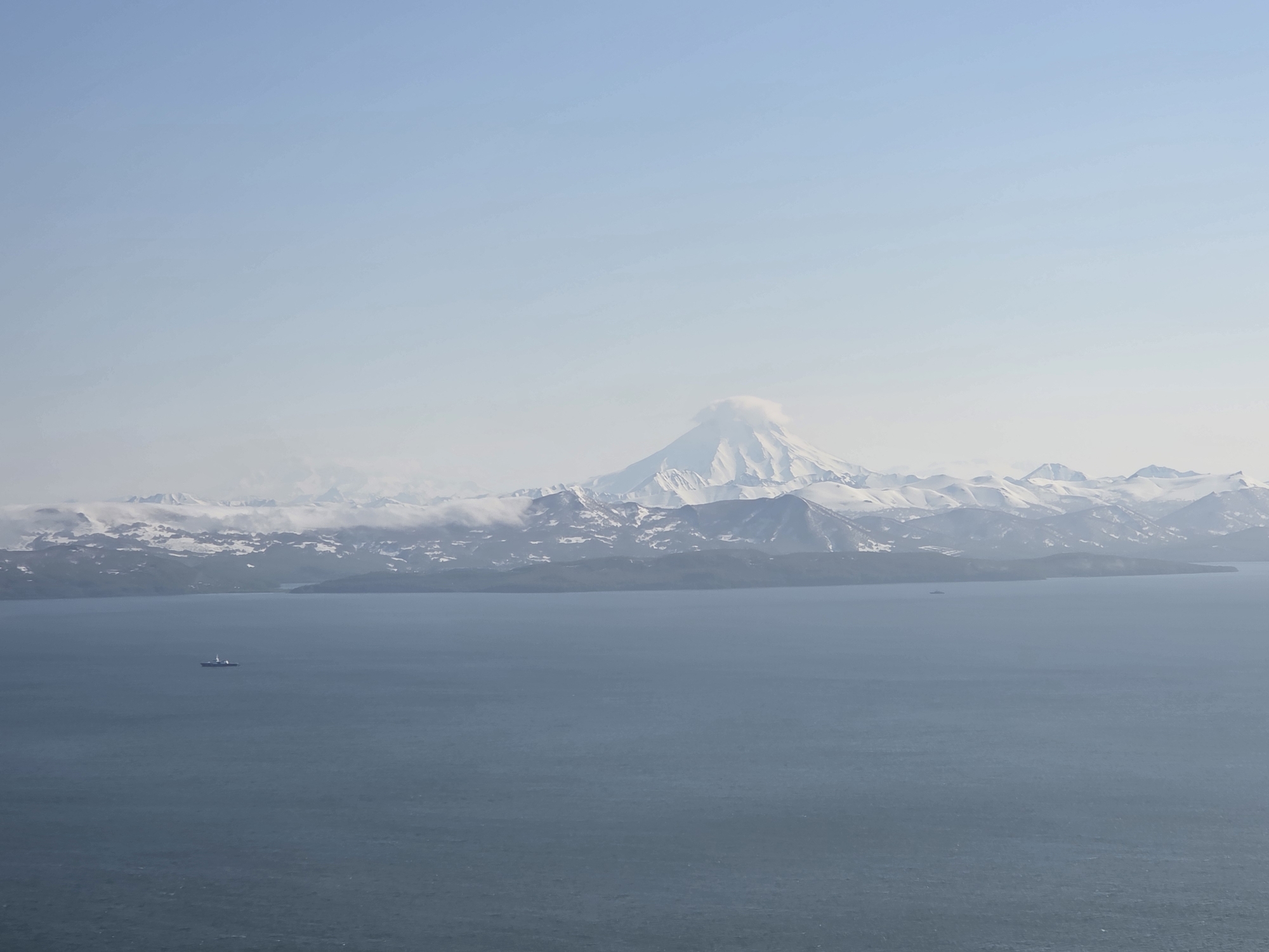 This is an aerial wide shot of a vast expanse of calm, dark blue ocean in the foreground. In the mid-ground, a body of land is visible, characterized by rugged, snow-capped mountains that stretch across the horizon. A plume of white smoke or cloud emanates from the peak of a prominent, conical volcano in the center of the mountain range. The sky above is a pale, clear blue with a gradient fading to white near the horizon. A small, dark ship with a white superstructure is positioned in the ocean to the left of the frame. The location is identified as being near the city of Krutoberegovy, Russia.