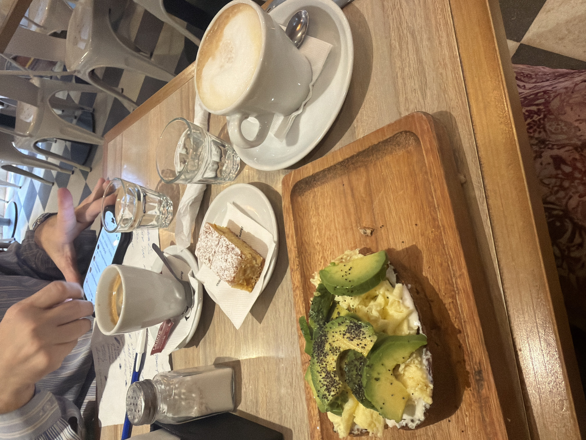 Here is a detailed description of the image:
**Location:** The image appears to have been taken in a café in Buenos Aires, Argentina. This is inferred from architectural or design elements or a reference point if present.
**Subjects:** The main subjects are a person and food/beverage items. The person is partially visible on the left, using a smartphone. The food and beverage items are arranged on a wooden table.
**Objects:**
* Two cups of coffee, one cappuccino and one espresso.
* A piece of cake on a small white plate.
* A wooden tray containing an open-faced sandwich topped with avocado and what appears to be scrambled egg.
* Two glasses of water
* A salt shaker
* A pen and some handwritten notes
* Plates and cutlery
* A smartphone
**Actions/Interactions:** The person is using their smartphone. The overall scene suggests someone is enjoying a meal or snack and potentially documenting it.
**Environment:** The environment is a café or restaurant setting. The table is wooden, and there are chairs visible. The area is well-lit, either naturally or with the use of overhead lighting.