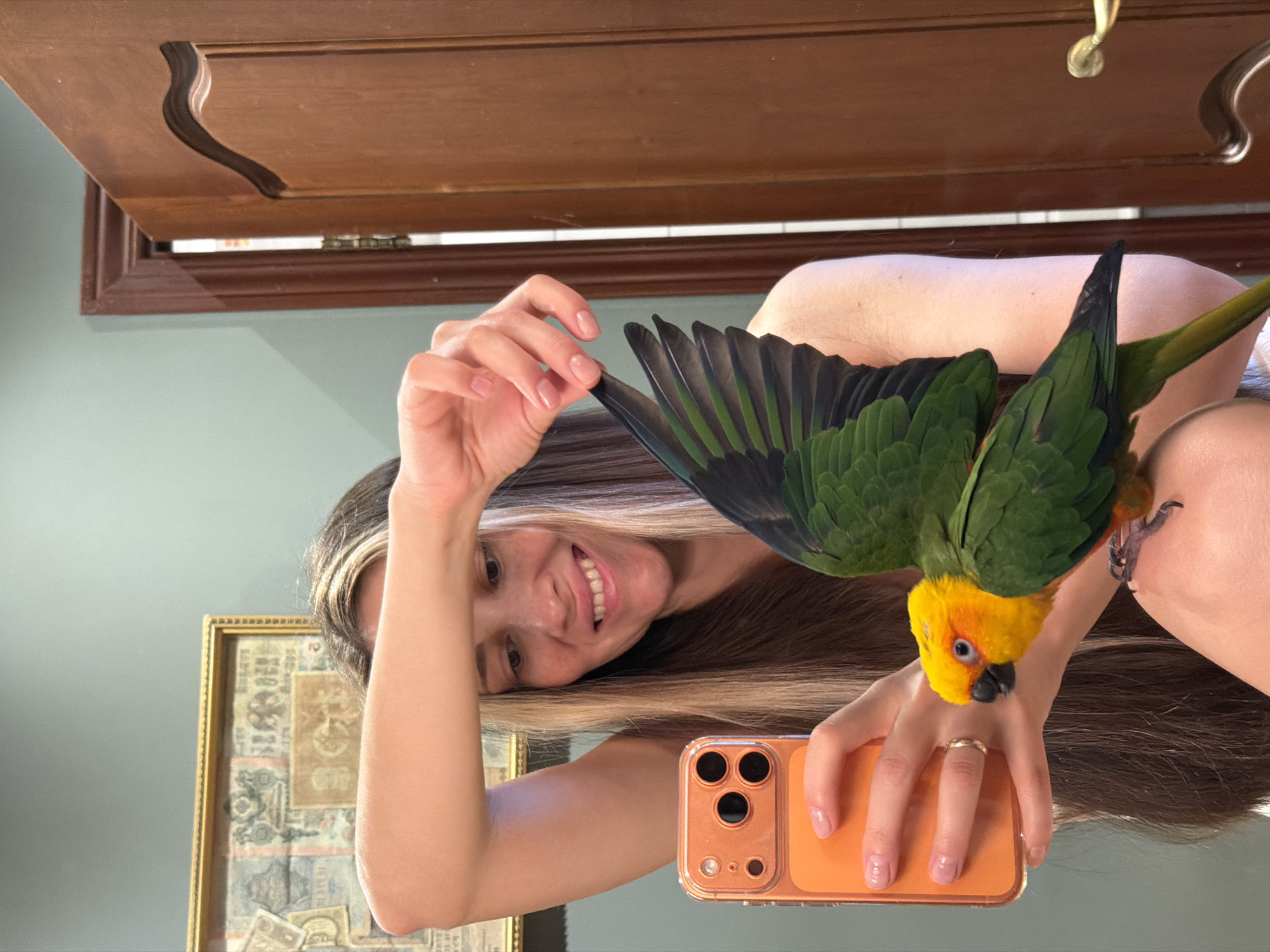 A fair-skinned woman with long brown and blonde hair is holding up a green and yellow parrot with black tail feathers. The woman is holding an orange-cased smartphone. The background includes a wall, a wooden cabinet, and a framed piece of art. This image doesn't indicate any location and does not provide information related to the city of Khimki, Russia.