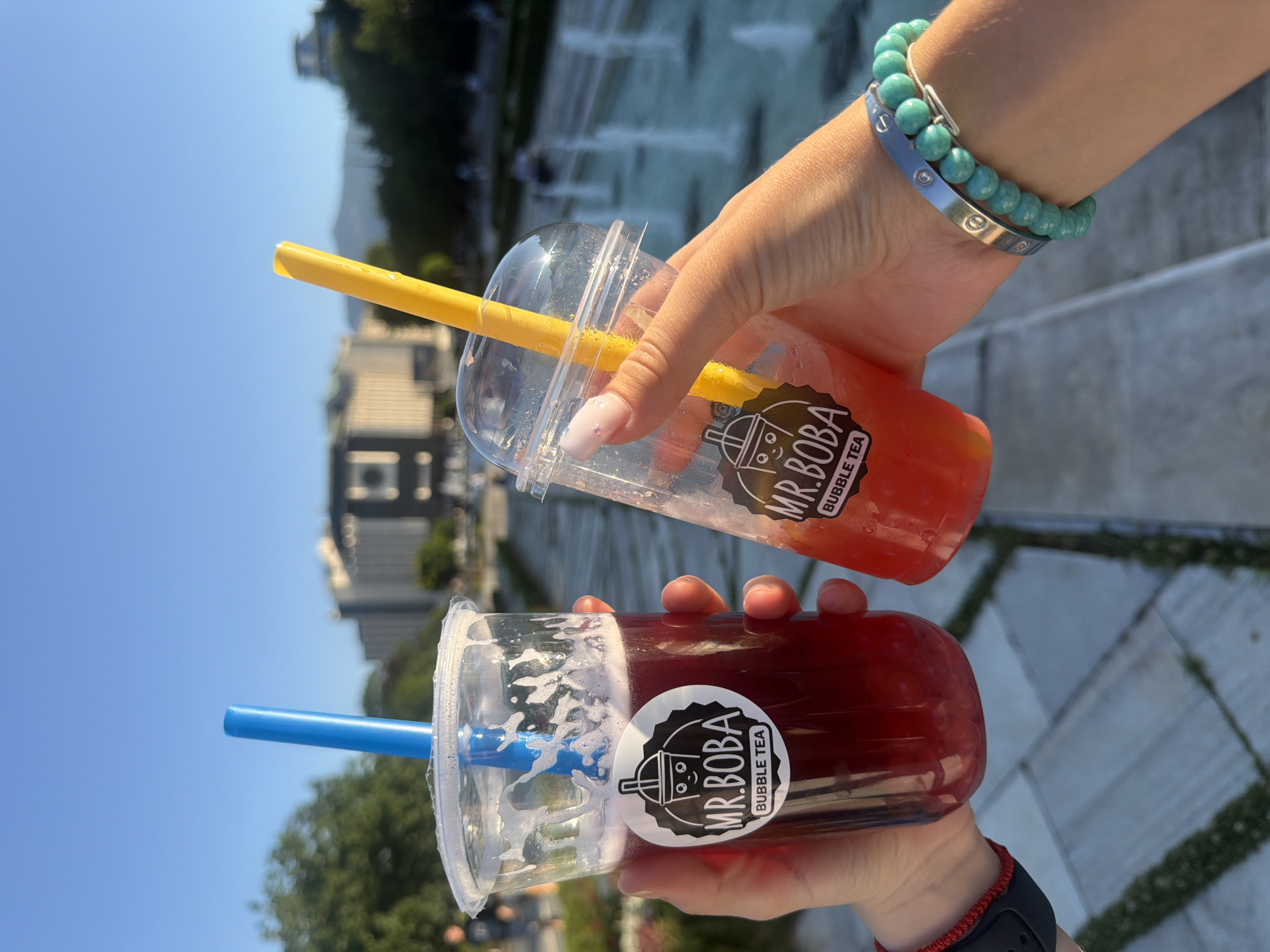 Two hands hold two "Mr. Boba Bubble Tea" cups against a clear blue sky. The cup on the left contains a dark red beverage with a blue straw, held by a hand with a red wristband. The cup on the right contains a reddish-orange beverage with a yellow straw, held by a hand with a white nail and a turquoise bracelet. In the background, a fountain is visible, suggesting the location is the area around the National Palace of Culture (NDK) in Sofia, Bulgaria, known for its park and fountains. The overall lighting indicates it is likely late afternoon or early evening.