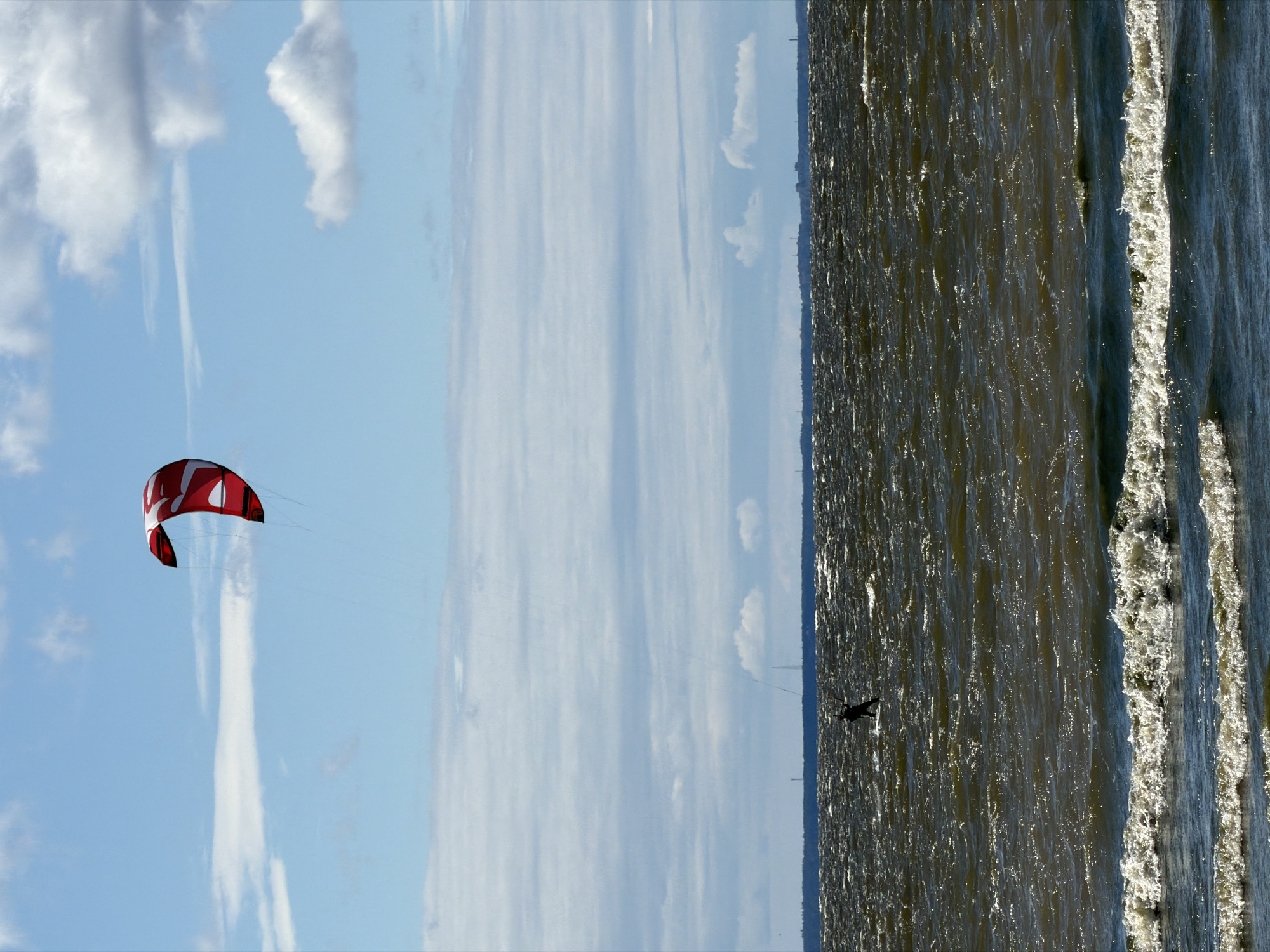 A single kiteboarder is in action on the choppy, dark waters of the Gulf of Finland, off the coast of Kronstadt, Russia. The kiteboarder, silhouetted against the waves, is positioned in the lower-left quadrant of the frame, facing right. Above, a large, red and white kite is prominent in the sky, tethered to the kiteboarder by thin lines. The sky is a mix of blue with scattered white clouds, some of which are thin and elongated, while others are more puffy. The horizon line, marking the distant shore of Kronstadt, is visible as a faint dark line with a few faint structures. The foreground of the image features white-capped waves breaking.