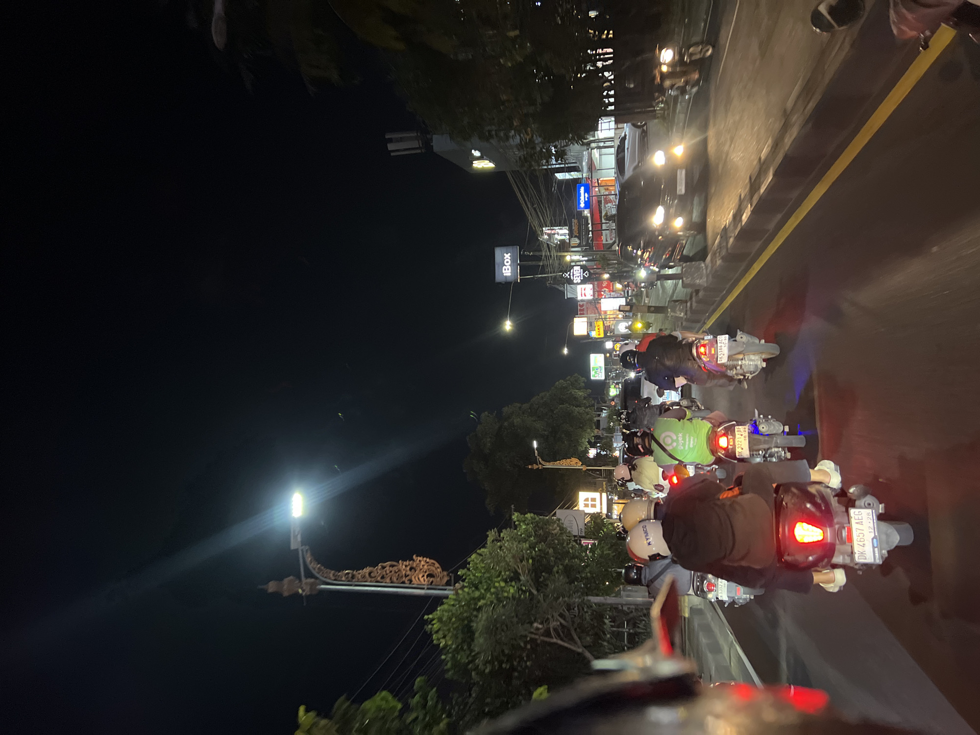 This image depicts a night scene on a busy street in Denpasar, Indonesia, likely during a period of traffic. Multiple motorbikes are the primary mode of transport visible, with riders wearing helmets. The motorcycles have their headlights and taillights illuminated. One motorbike in the foreground displays a license plate reading "DK 4657 AEG". Buildings with illuminated signs line the street, including "iBox" and other commercial establishments. Trees are present along the roadside, and streetlights illuminate the scene. The road surface is paved with a yellow line marking the lane. The atmosphere suggests a typical urban environment at night, with active traffic and commercial activity.