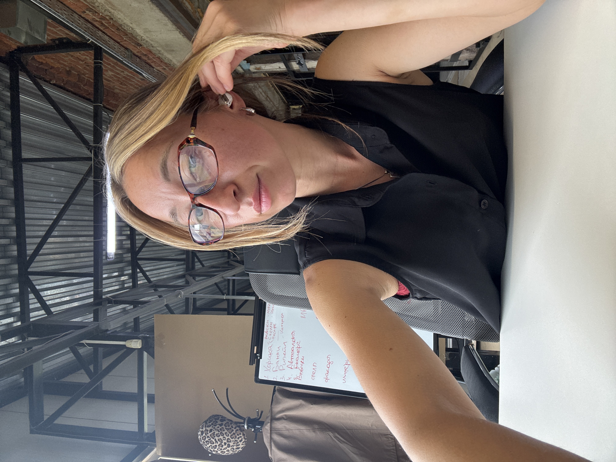 This is a vertical, waist-up, selfie-style photo of a woman in an office or workshop setting. The woman has blonde hair and is wearing glasses with brown frames and a black shirt. She is looking towards the camera with a neutral expression. Her left hand is resting on her head, and her right arm is bent, with her forearm visible in the foreground.
Behind the woman, a whiteboard is visible with handwritten text in Cyrillic. The text appears to be a list of items, possibly project tasks or categories. The background shows industrial-style architecture, with exposed brickwork, metal beams, and corrugated metal sheeting, suggesting a loft-like or open-plan workspace. A coat rack with a leopard-print hat is also visible in the lower left corner of the image. The lighting suggests it is daytime. The overall impression is that of a candid moment during a workday.