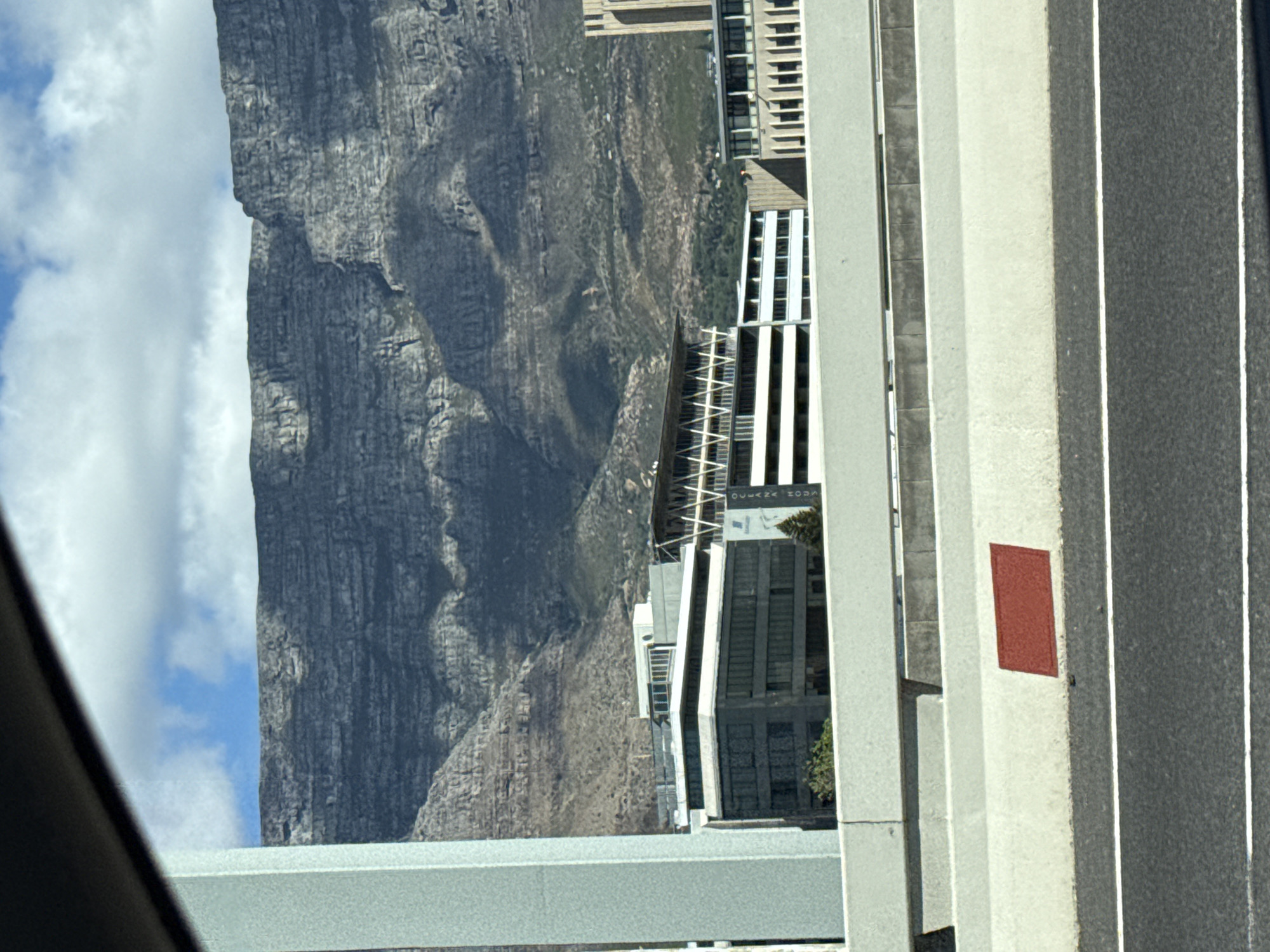 The image shows a view of Table Mountain in Cape Town, South Africa, taken from a vehicle or elevated position. The mountain dominates the left side of the frame, with visible cloud cover. In the foreground, a concrete highway barrier with a small rectangular red marker is seen, indicating a road. Modern buildings are situated between the mountain and the highway.