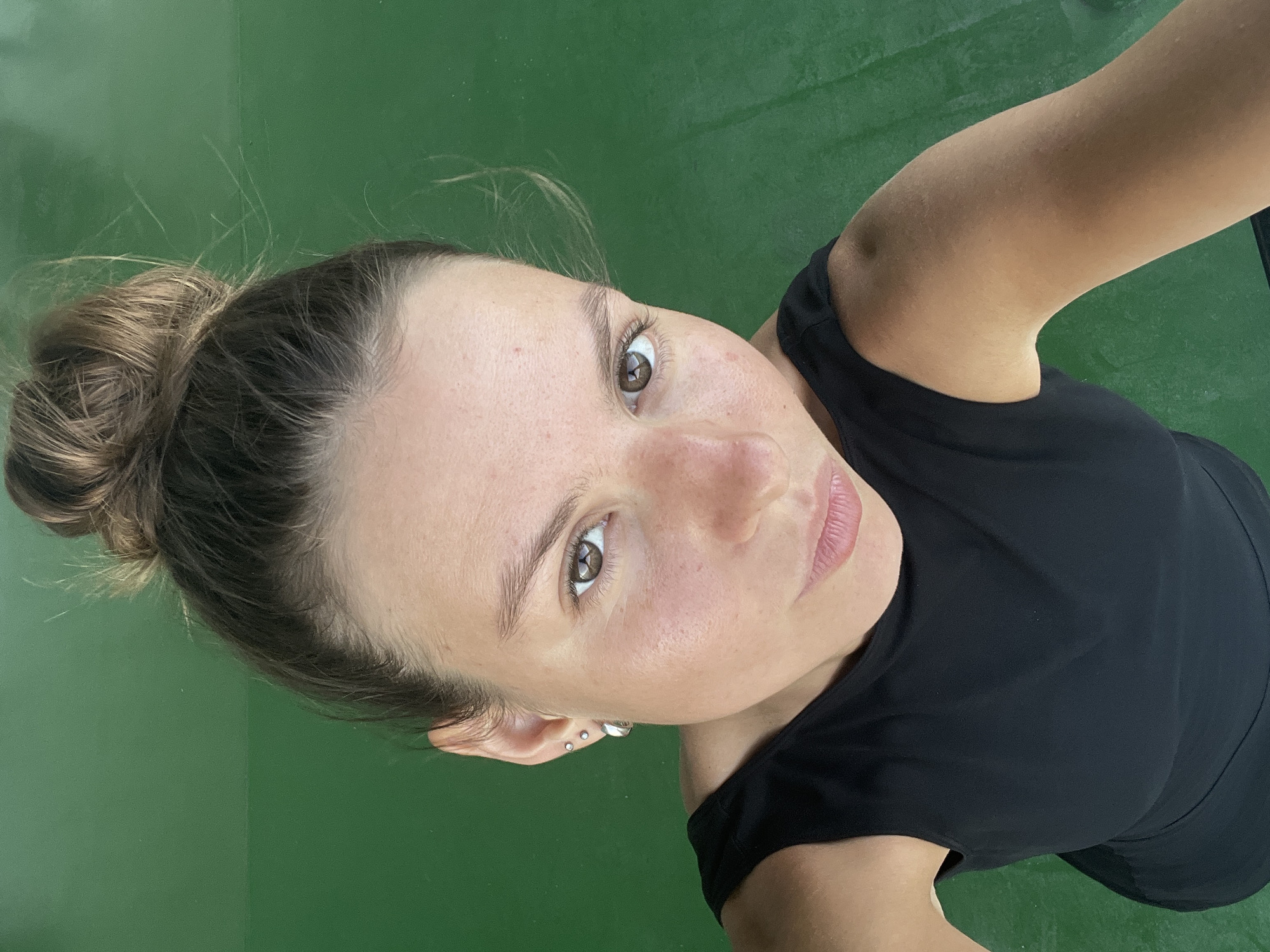 A young woman with her hair in a bun is looking up at the camera. She is wearing a black top and has several earrings in her left ear. The background is a solid green surface. The image appears to be a selfie taken outdoors. The location is not discernible from the image itself and is therefore not included.