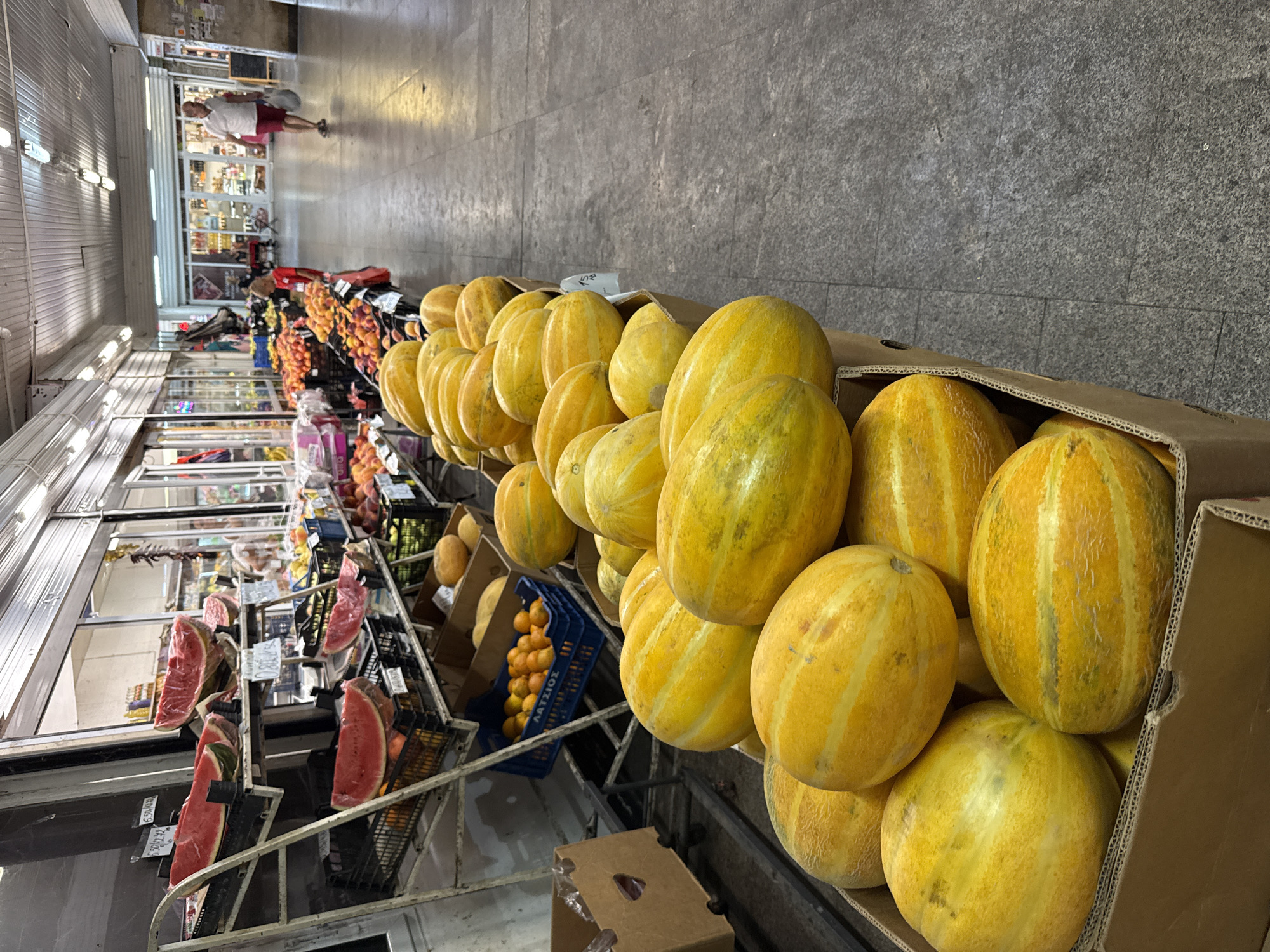 The image depicts a fruit stall within a covered market, likely located in Burgas, Bulgaria, given the presence of Greek signage ("ΛΑΤΣΙΟΣ" appears on a blue crate). The foreground is dominated by several cardboard boxes filled with ripe yellow and green striped melons. Behind these, on tiered metal shelves, are more fruits including what appear to be sliced watermelons, oranges, and peaches. A person, a young boy in a red shirt, stands near the fruit display, looking towards the left. Further down the market aisle, a man in a white shirt and red shorts walks away from the camera, carrying a white bag. The market environment features a tiled floor, a suspended ceiling with lighting, and storefronts lining both sides of the aisle. The overall scene suggests a bustling marketplace filled with fresh produce.