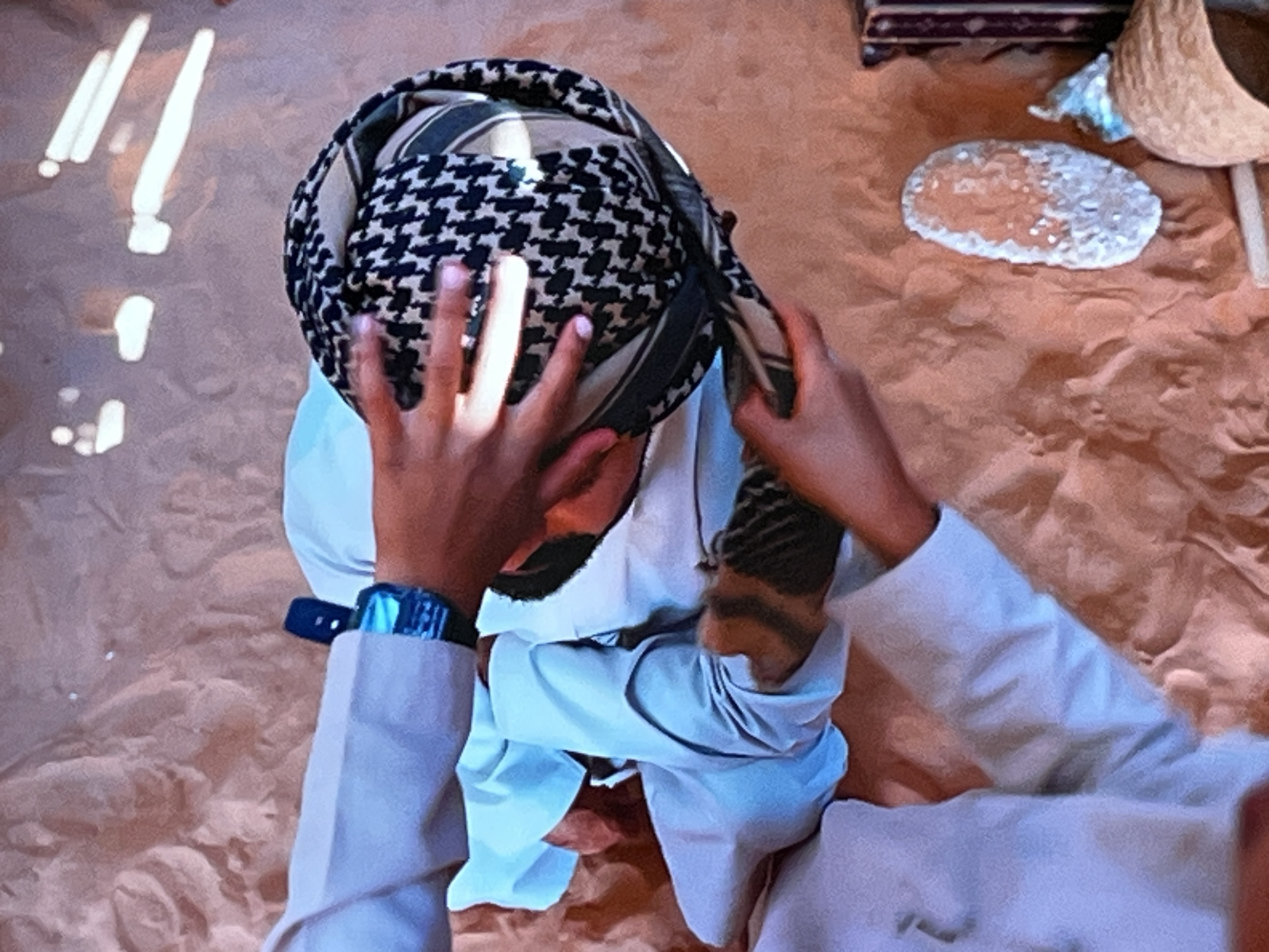 The image shows a person in a desert-like environment, possibly in Kharkiv, Ukraine, adjusting a keffiyeh, a traditional Middle Eastern headdress. The person is wearing a light-colored robe and a dark-strapped watch on their left wrist. In the background, there are elements suggestive of a temporary setup: a woven basket, a flat, round object that appears to be bread, and a striped fabric. The ground is sandy and uneven. The lighting in the image is strong, casting noticeable shadows.