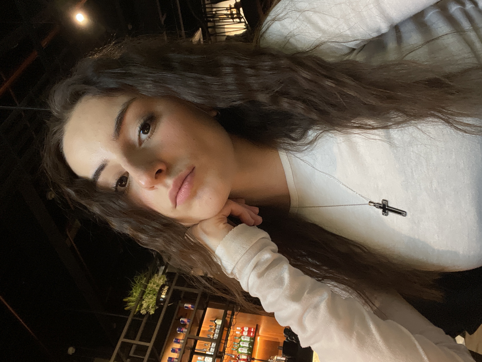 A young woman with long, wavy brown hair is the primary subject of the image. She is wearing a light-colored, long-sleeved shirt and a silver necklace with a cross pendant. Her head rests on her hand, and she gazes towards the camera.
The location is inside a bar or restaurant in Moscow, Russia. Shelves filled with alcohol bottles are visible in the background, including what appears to be Red Bull cans and wine bottles. A sign reads "Bruni." The scene is dimly lit, with artificial lighting illuminating parts of the background.